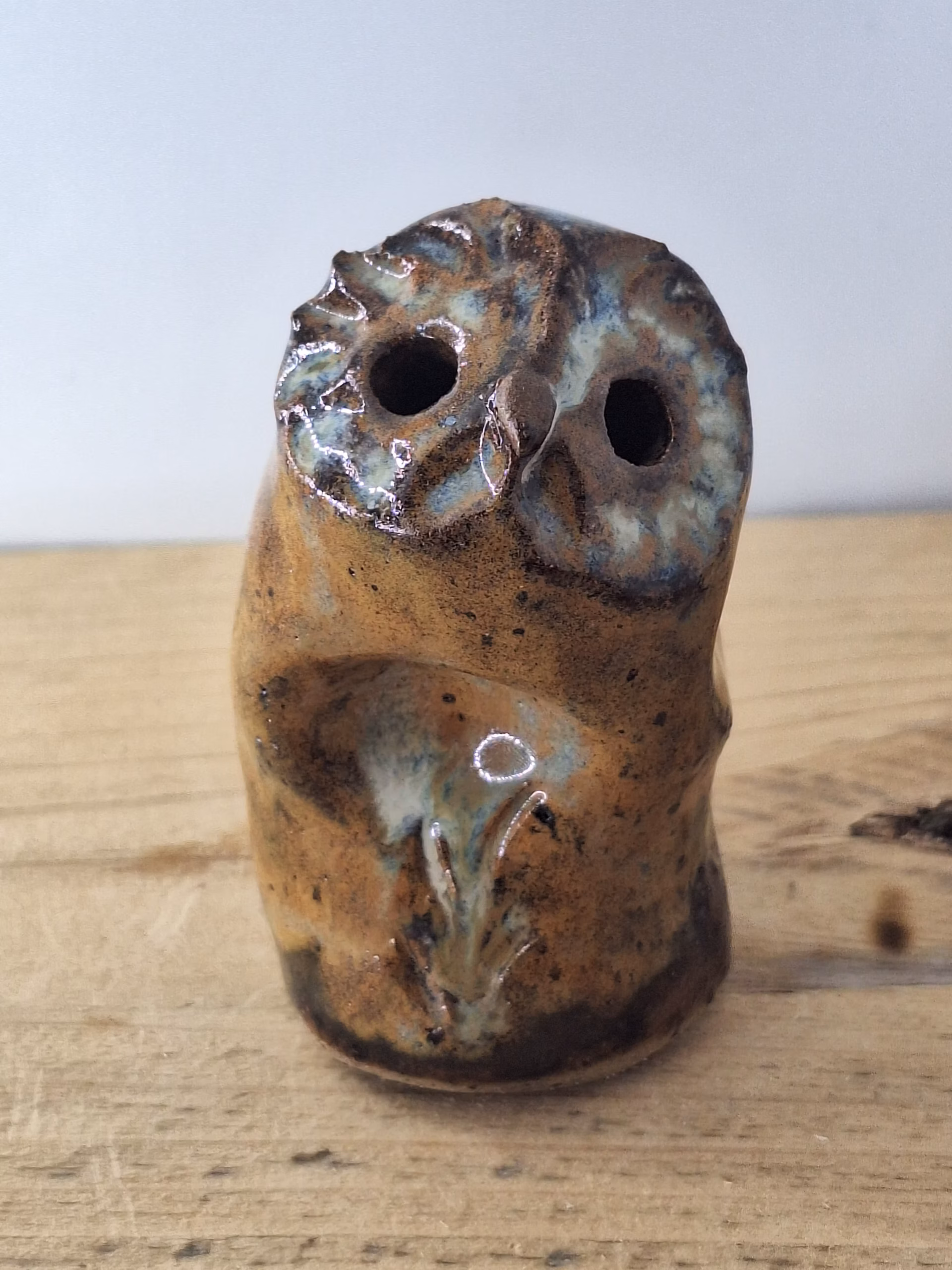 Small ceramic owl figurine with mottled brown and blue glaze, round head with large circular eyes and textured surface.