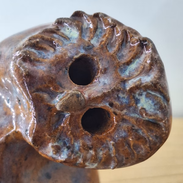 Small ceramic owl with textured feather detail, warm brown tones and blue-grey accents, two round eye holes.