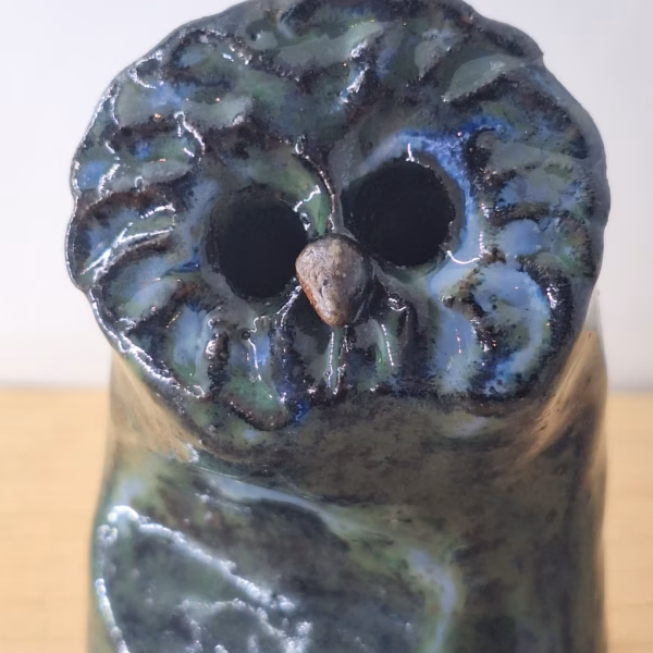 Small ceramic owl with large round eyes, mottled blue and green surface, and a tiny spider perched on its chest.