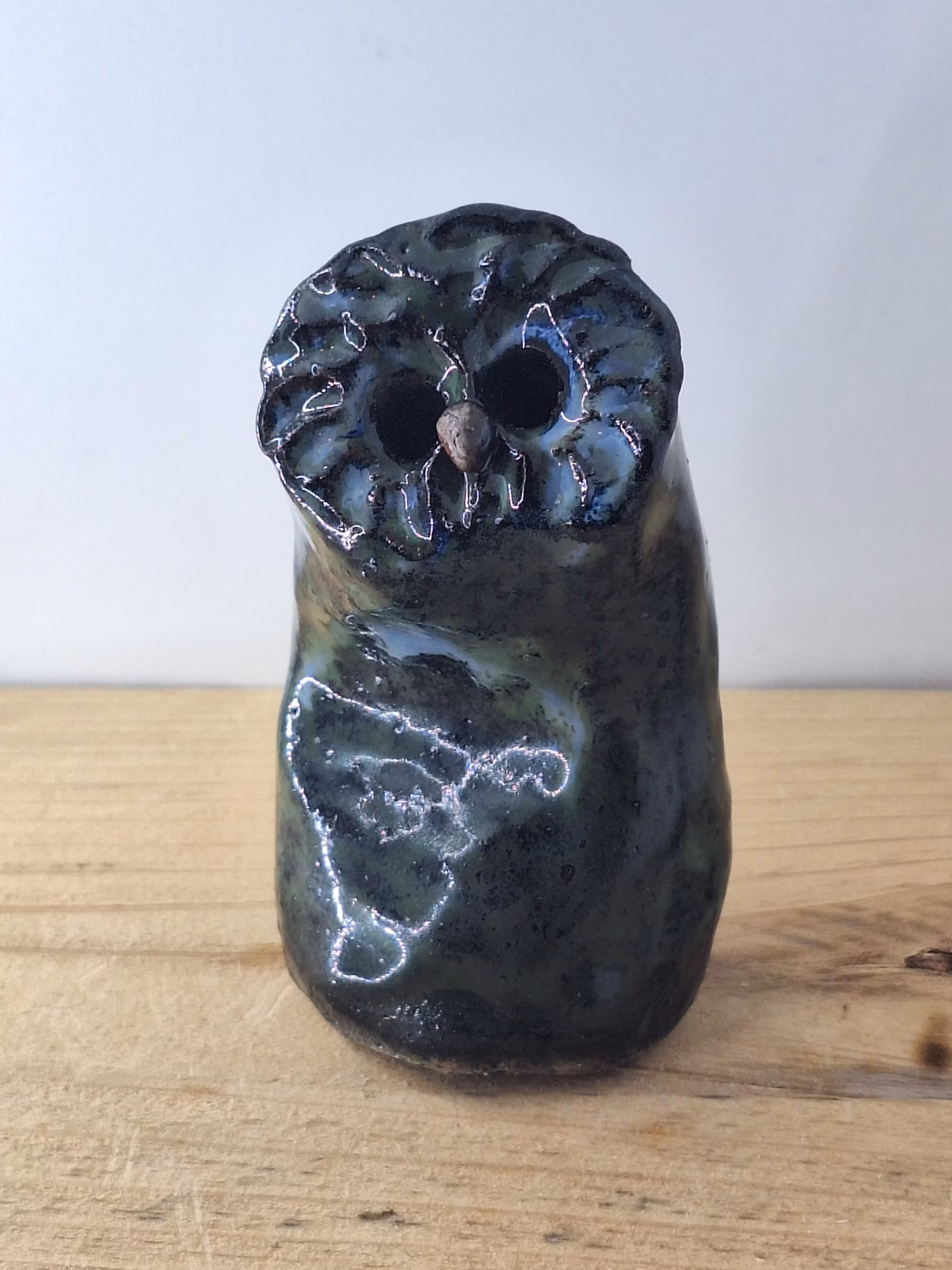 Small ceramic owl with large round eyes, mottled blue and green surface, and a tiny spider perched on its chest.