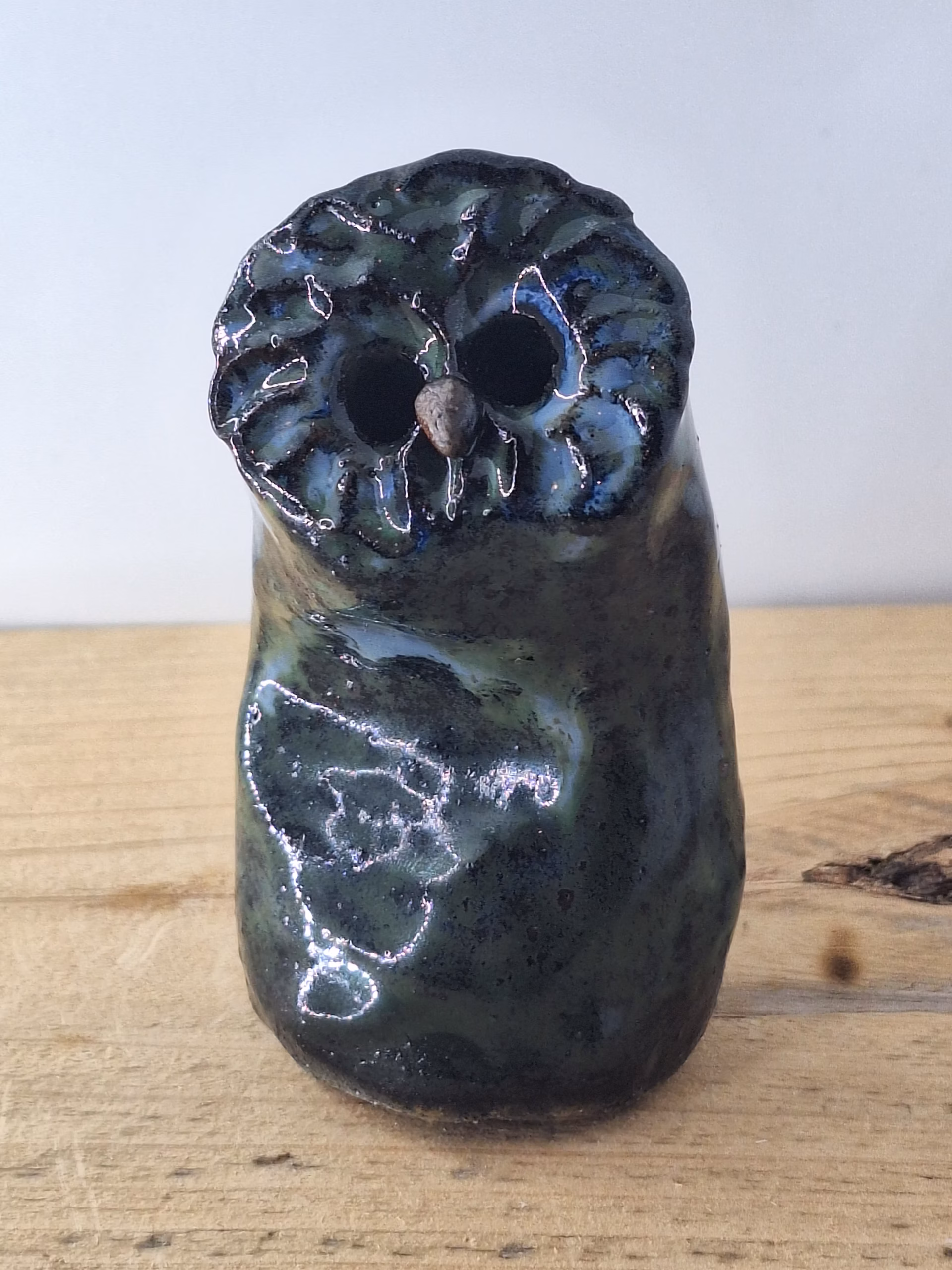 Small ceramic owl with large round eyes, mottled blue and green surface, and a tiny spider perched on its chest.
