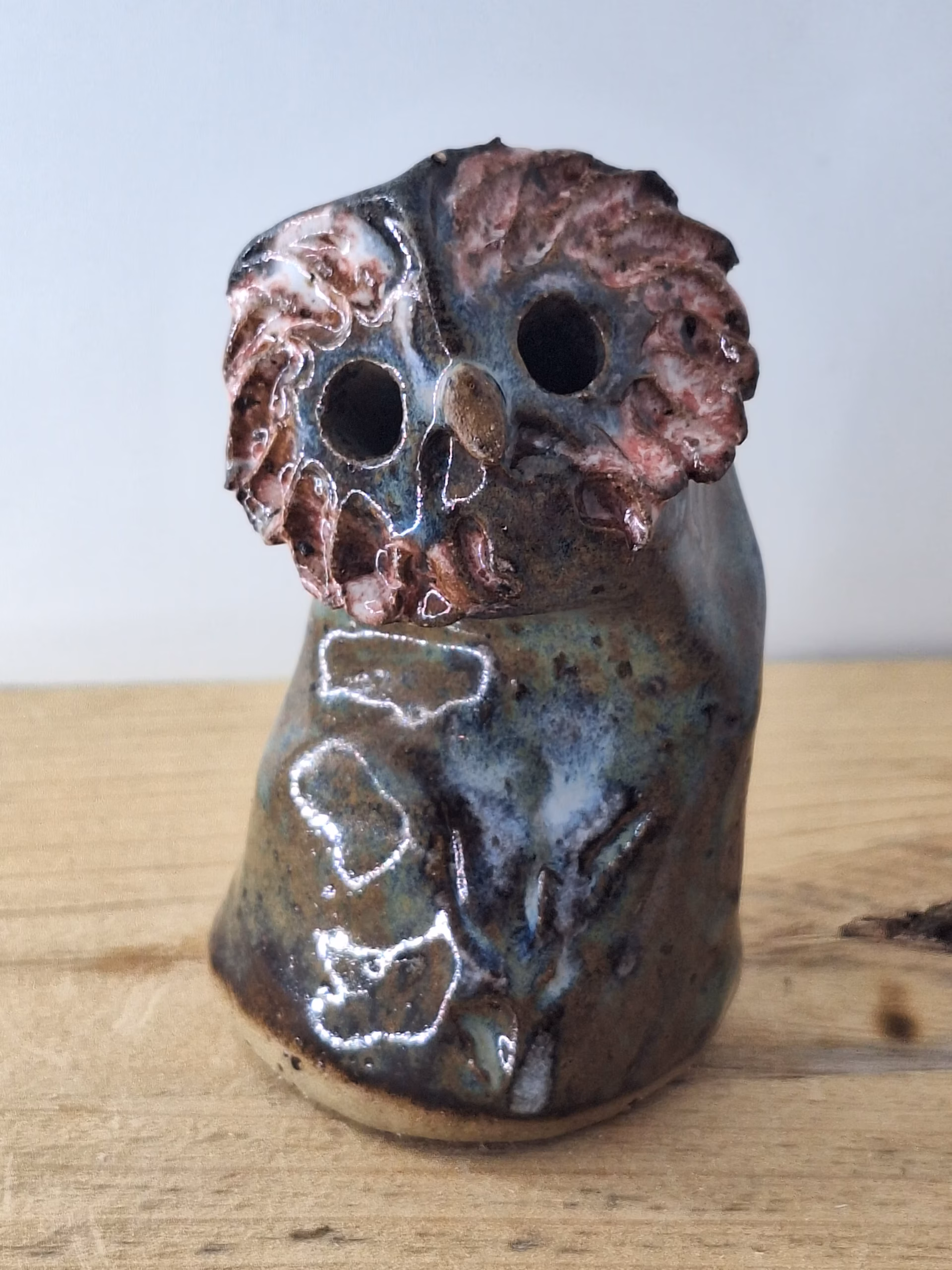 small ceramic owl head with textured pink and blue surface, dark round eyes, perched on a cylindrical base.