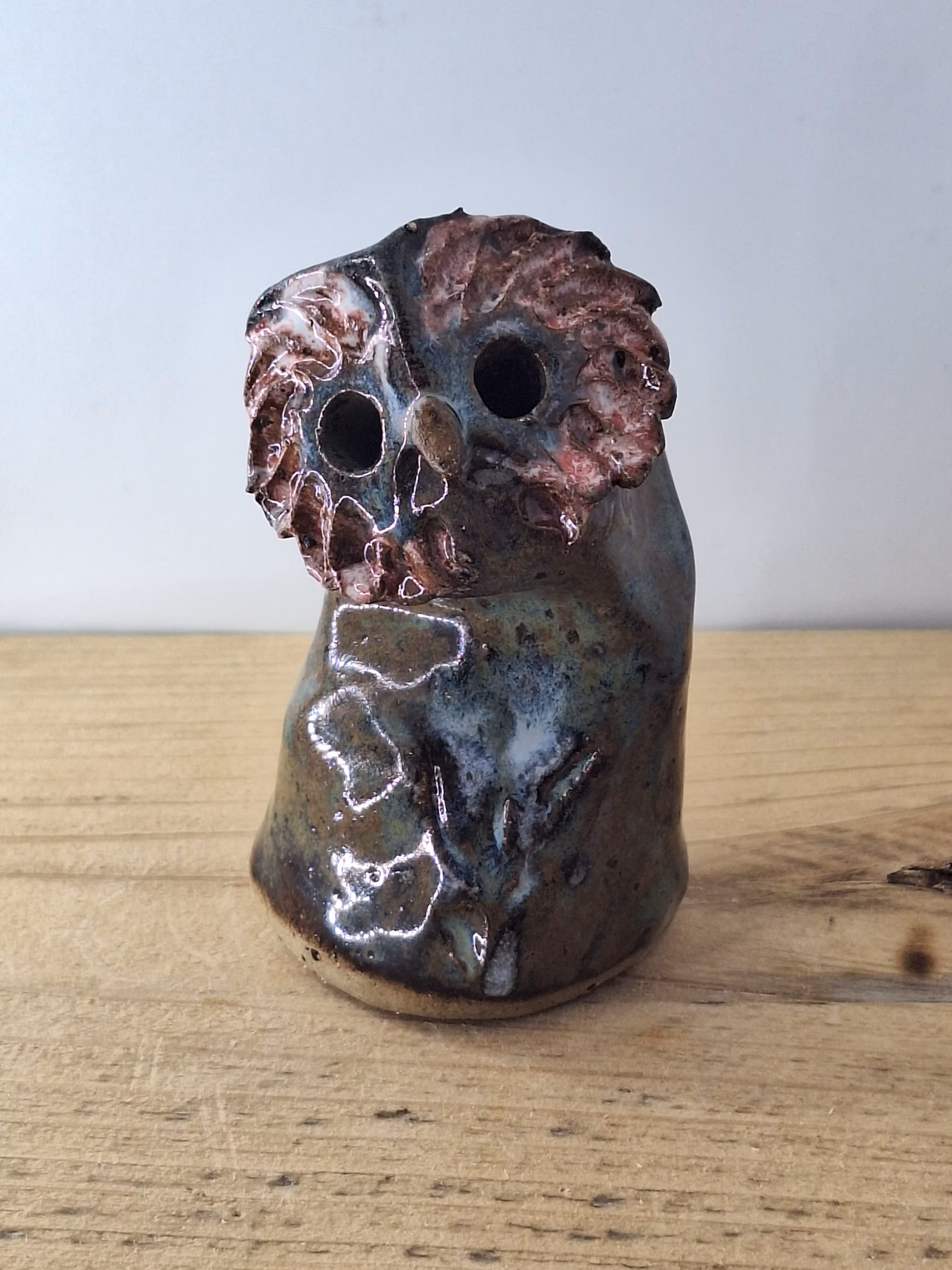small ceramic owl head with textured pink and blue surface, dark round eyes, perched on a cylindrical base.