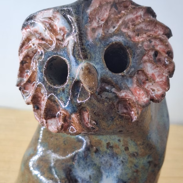 small ceramic owl head with textured pink and blue surface, dark round eyes, perched on a cylindrical base.