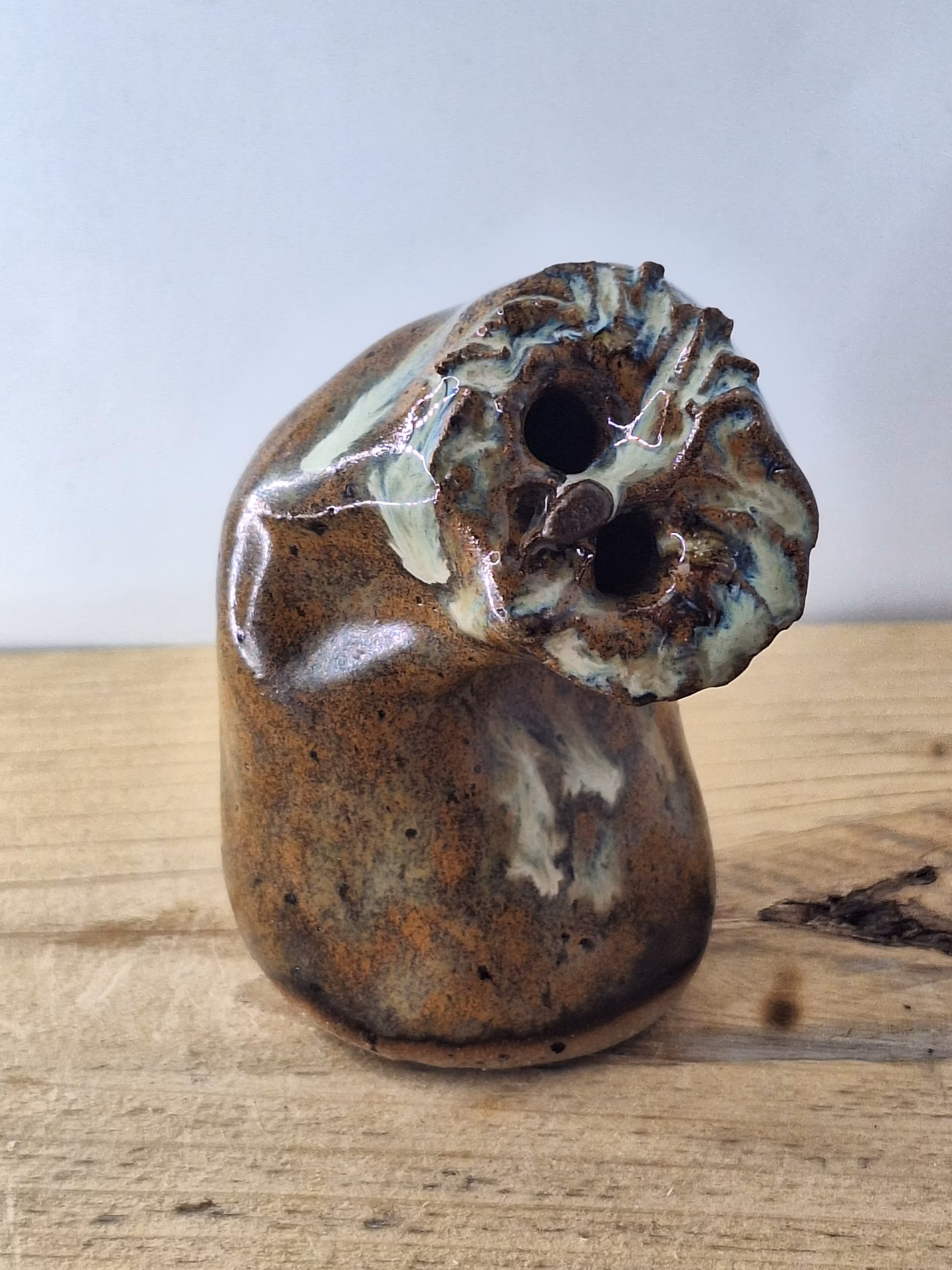 Small ceramic owl with a rounded body, textured surface in warm browns and creams, two dark hollow eyes, and a contemplative expression.