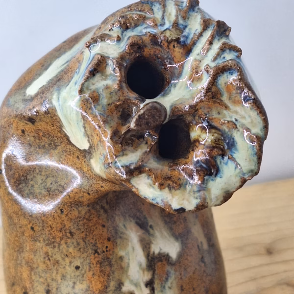 Small ceramic owl with a rounded body, textured surface in warm browns and creams, two dark hollow eyes, and a contemplative expression.