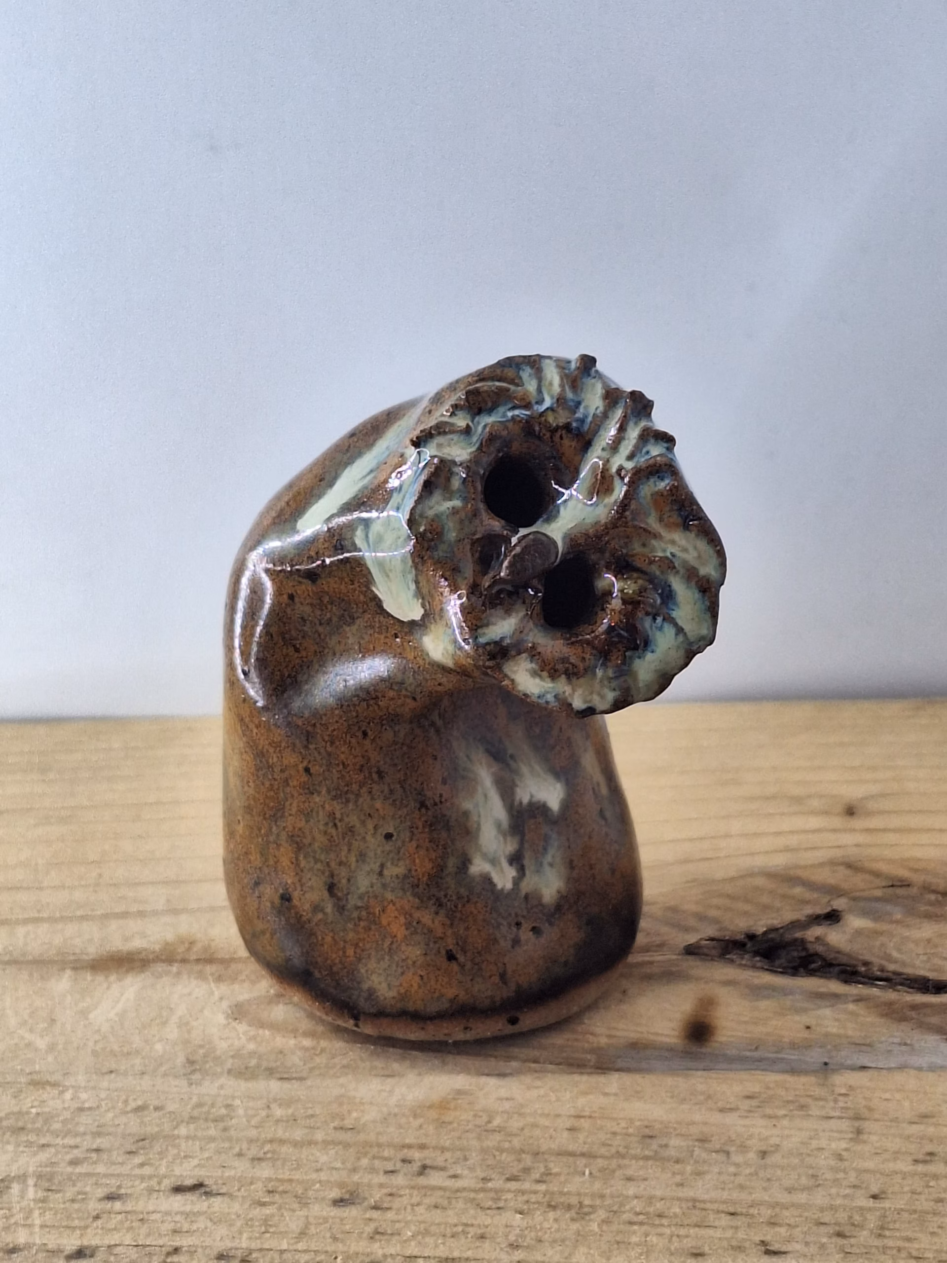 Small ceramic owl with a rounded body, textured surface in warm browns and creams, two dark hollow eyes, and a contemplative expression.