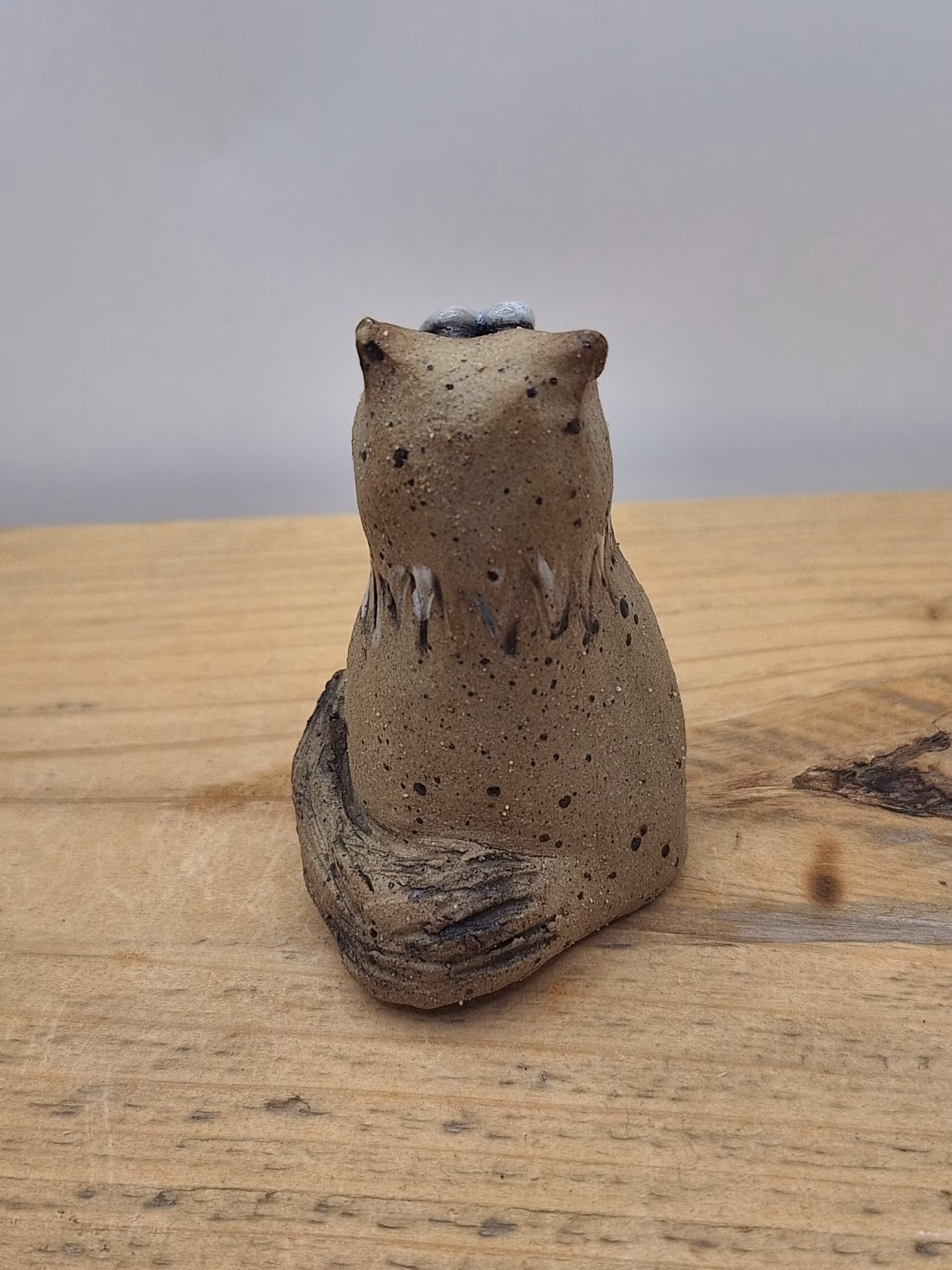 Small ceramic cat figurine with wide-set eyes, textured brown surface, and painted white markings on chest and face.