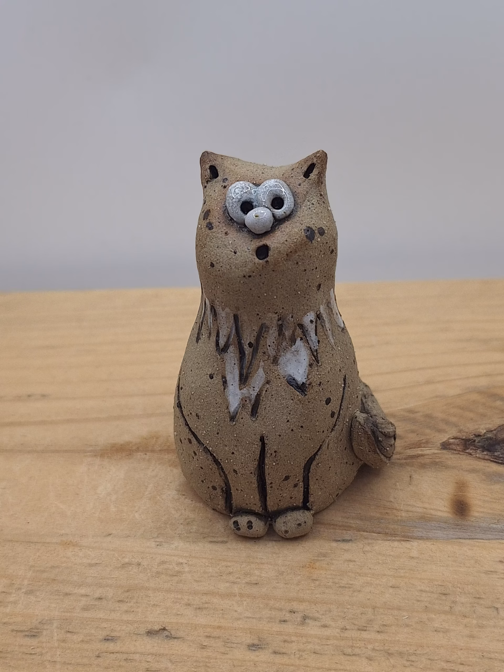 Small ceramic cat figurine with wide-set eyes, textured brown surface, and painted white markings on chest and face.
