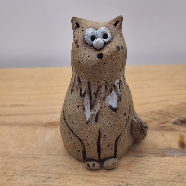Small ceramic cat figurine with wide-set eyes, textured brown surface, and painted white markings on chest and face.