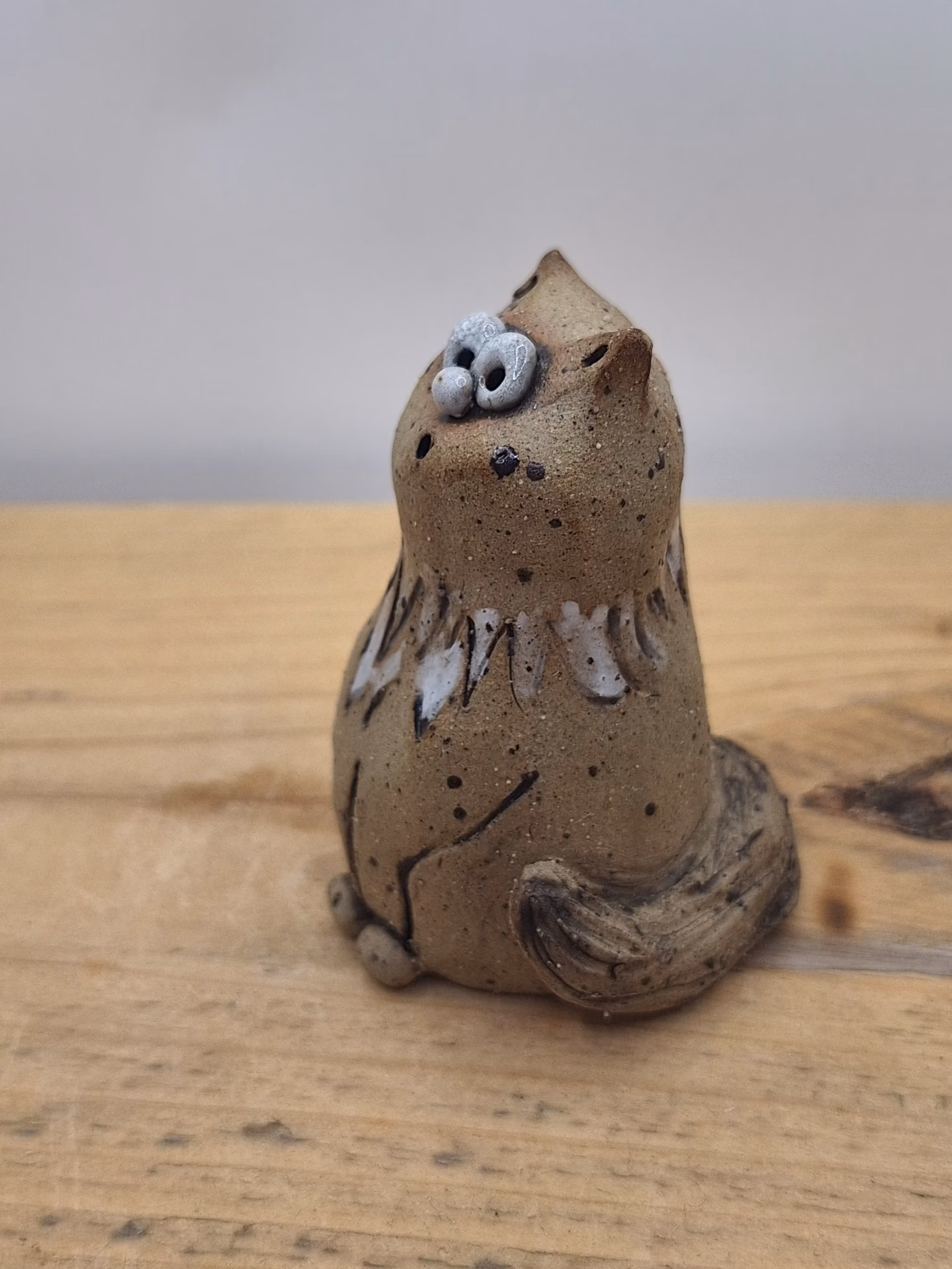 Small ceramic cat figurine with wide-set eyes, textured brown surface, and painted white markings on chest and face.
