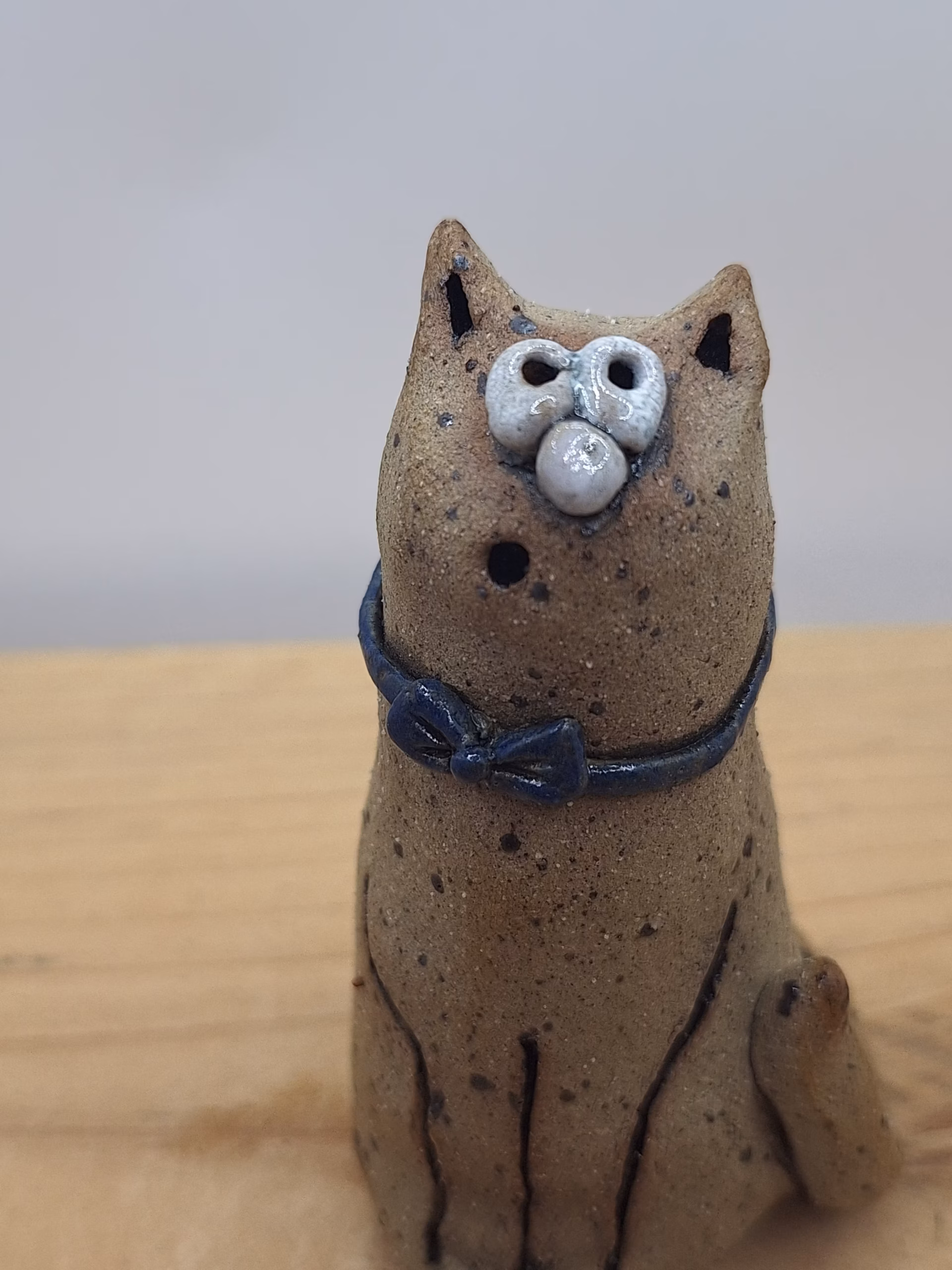 Small ceramic cat figurine with textured sandy surface, white painted face with wide eyes, pointed ears, and a dark blue bow tie around its neck.