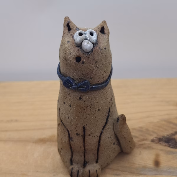 Small ceramic cat figurine with textured sandy surface, white painted face with wide eyes, pointed ears, and a dark blue bow tie around its neck.