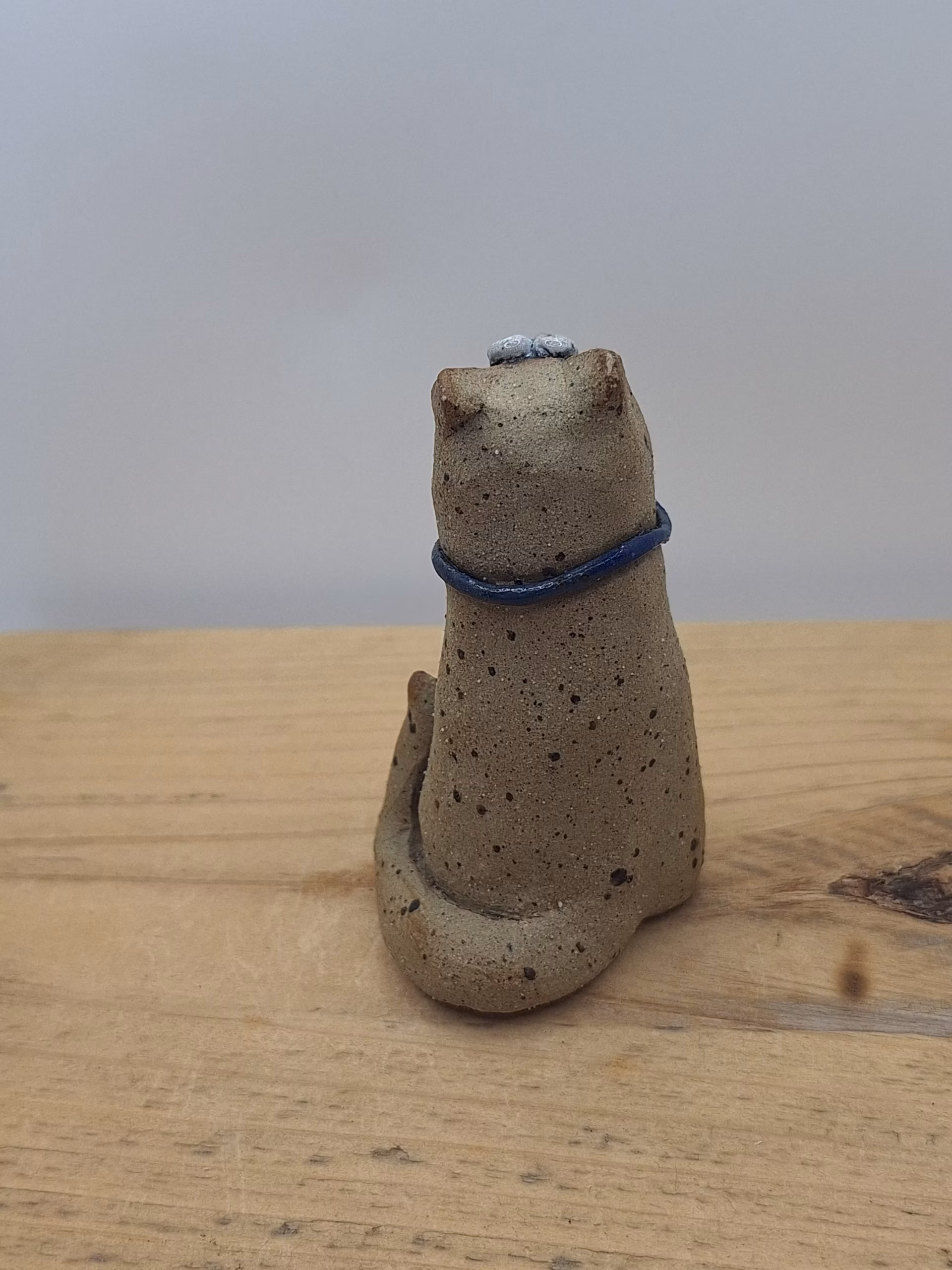 Small ceramic cat figurine with textured sandy surface, white painted face with wide eyes, pointed ears, and a dark blue bow tie around its neck.