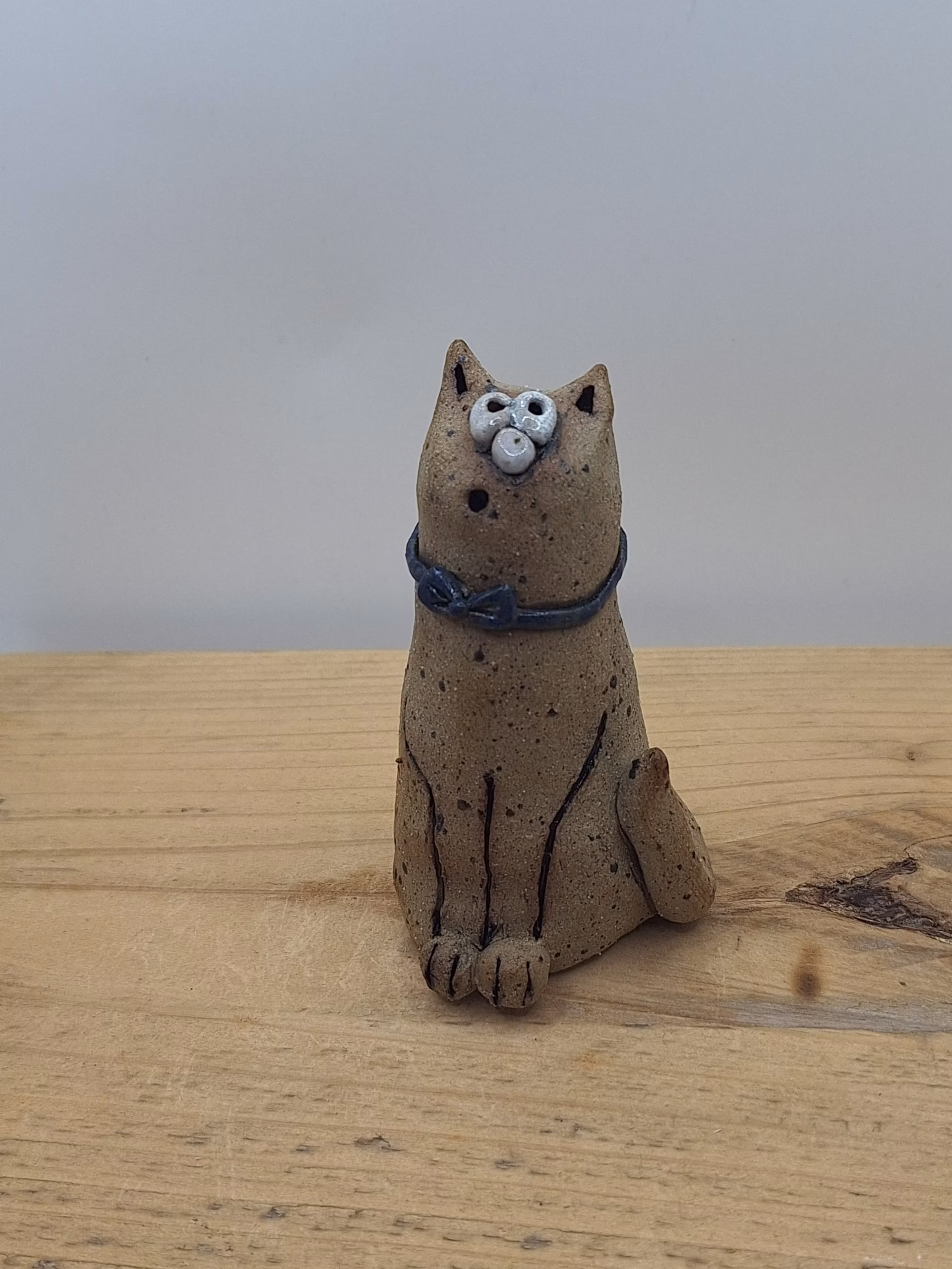 Small ceramic cat figurine with textured sandy surface, white painted face with wide eyes, pointed ears, and a dark blue bow tie around its neck.