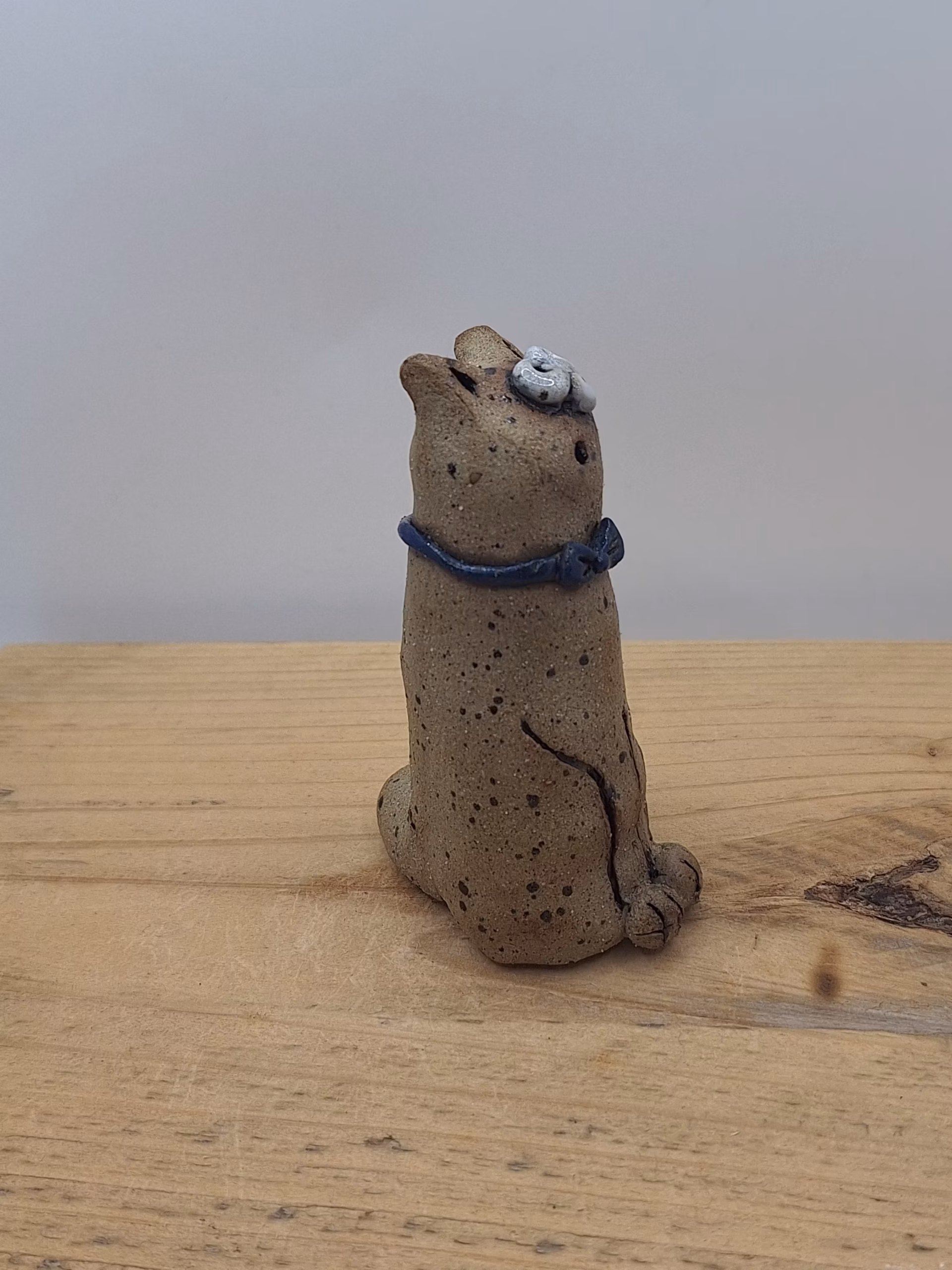 Small ceramic cat figurine with textured sandy surface, white painted face with wide eyes, pointed ears, and a dark blue bow tie around its neck.