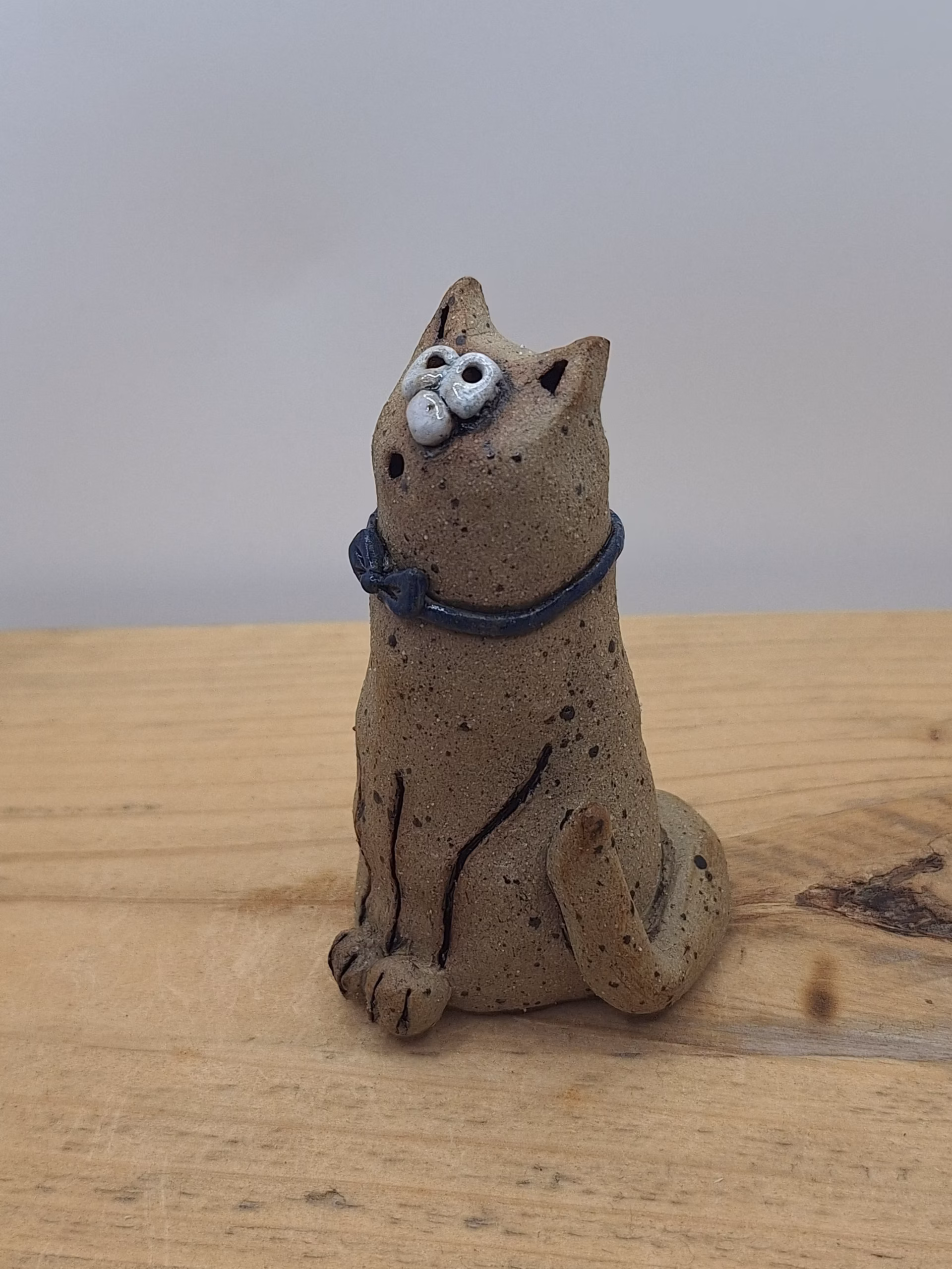 Small ceramic cat figurine with textured sandy surface, white painted face with wide eyes, pointed ears, and a dark blue bow tie around its neck.
