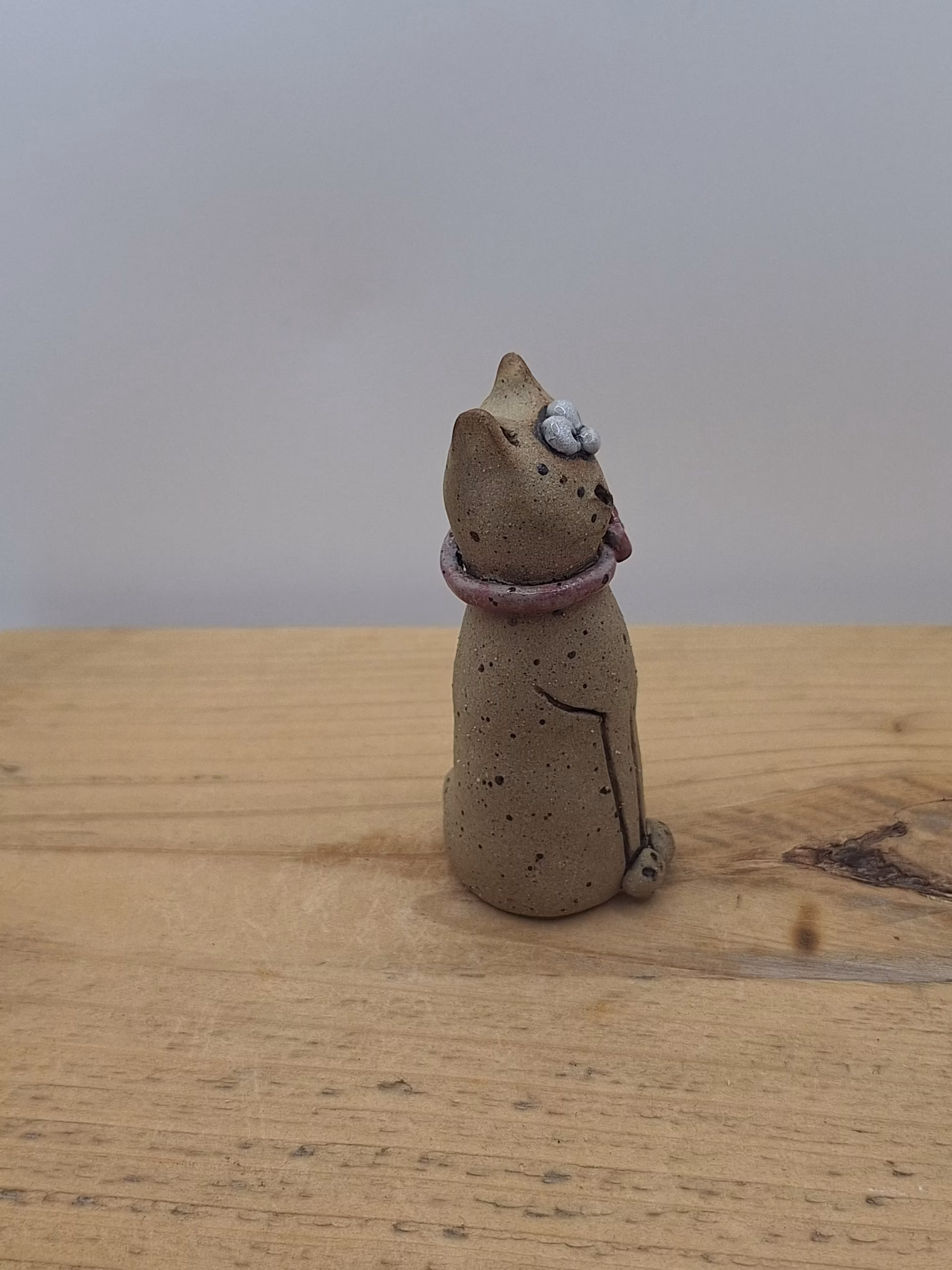 Small ceramic cat figurine with wide eyes, speckled body, pink bow collar, and one raised paw.