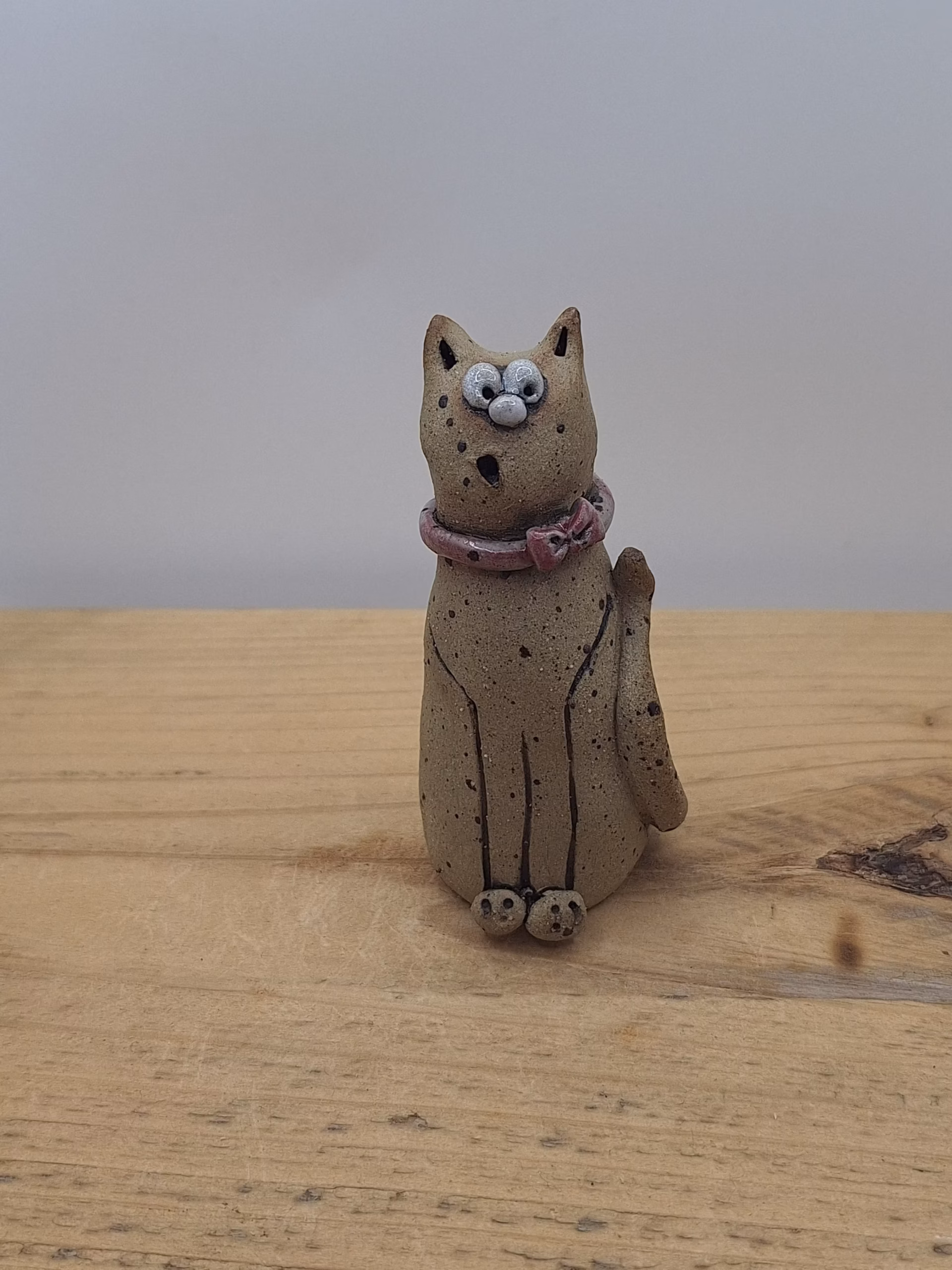 Small ceramic cat figurine with wide eyes, speckled body, pink bow collar, and one raised paw.