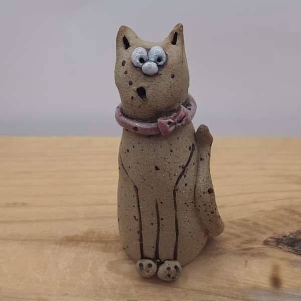Small ceramic cat figurine with wide eyes, speckled body, pink bow collar, and one raised paw.