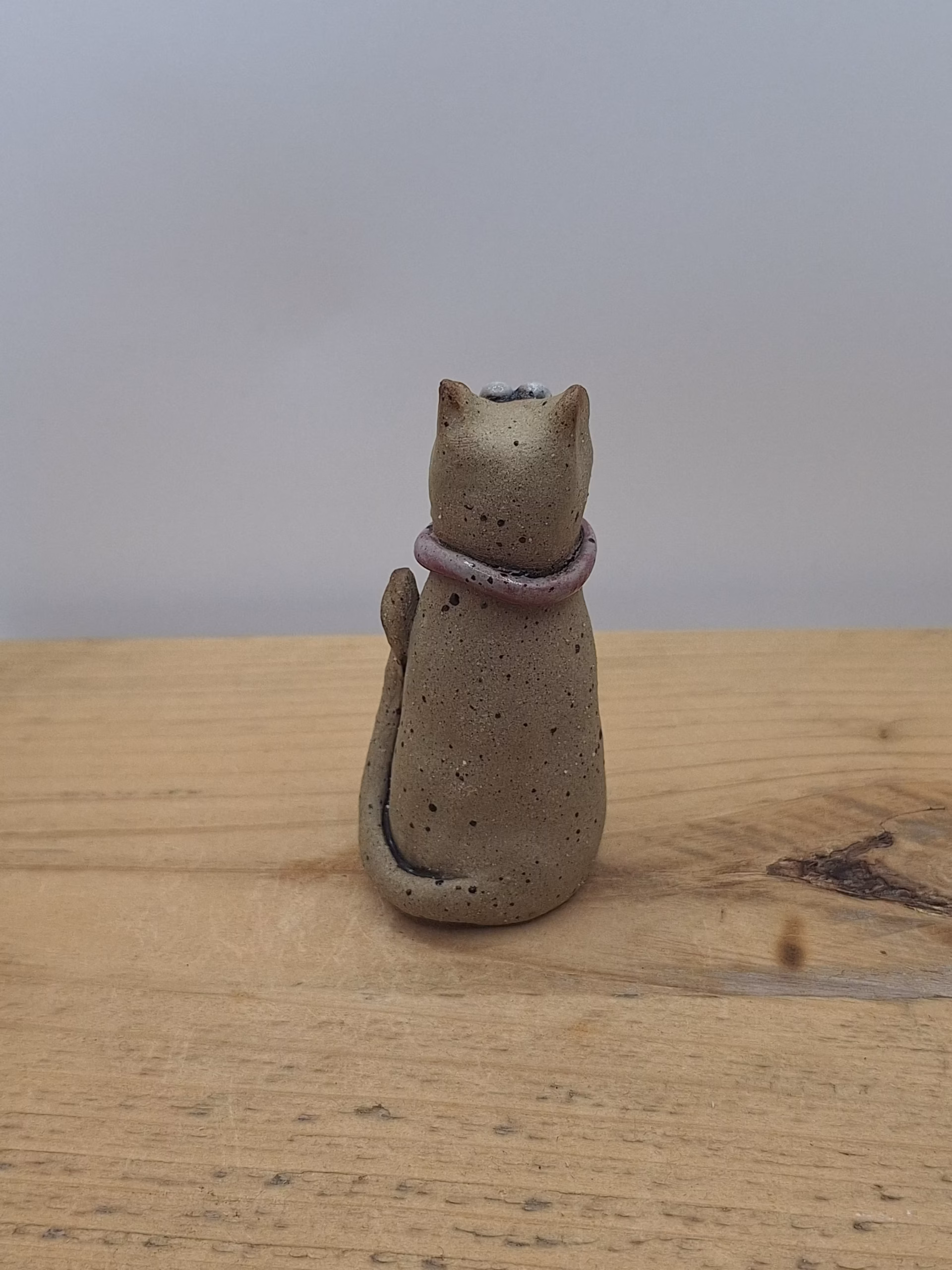 Small ceramic cat figurine with wide eyes, speckled body, pink bow collar, and one raised paw.