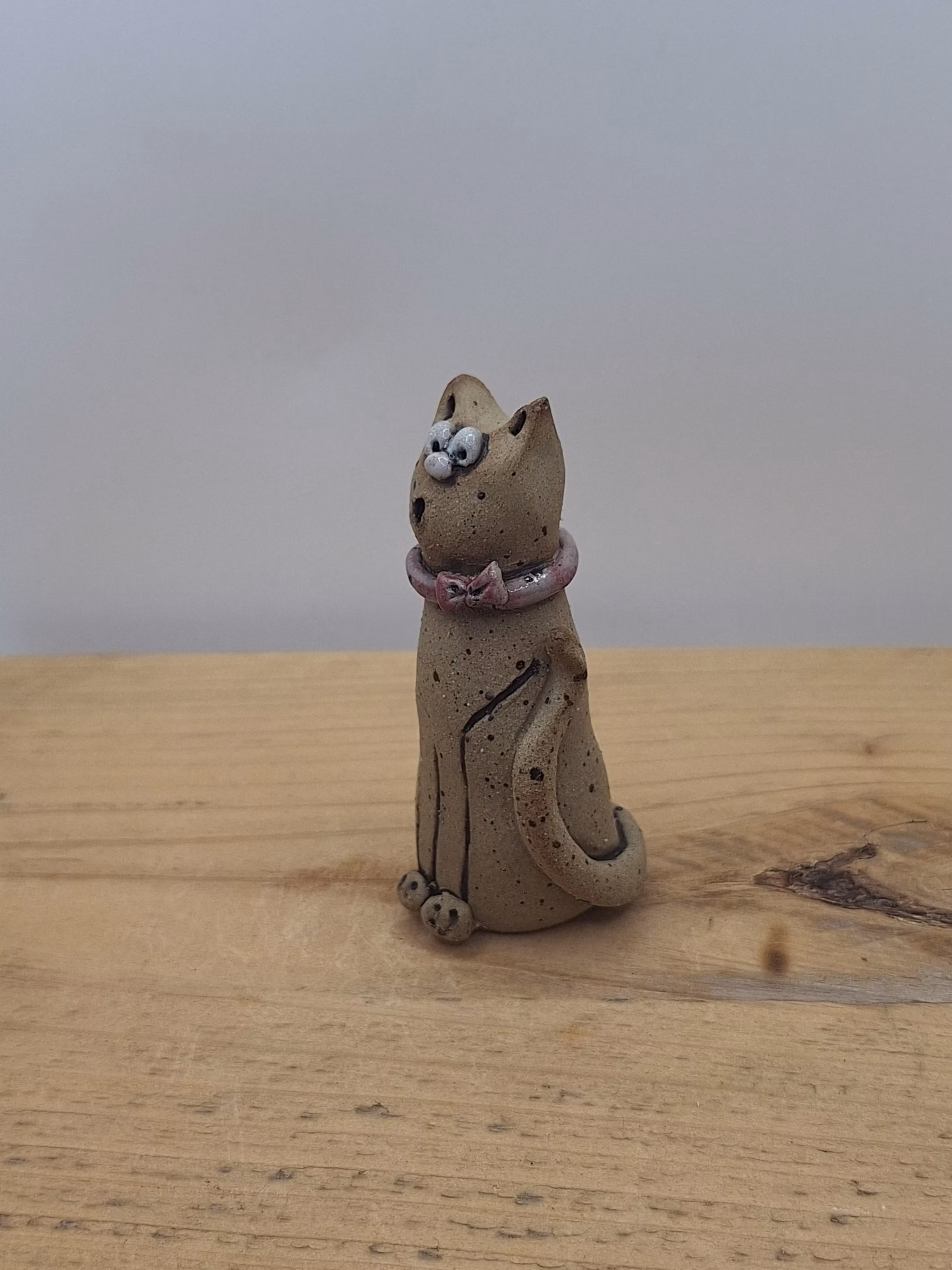 Small ceramic cat figurine with wide eyes, speckled body, pink bow collar, and one raised paw.