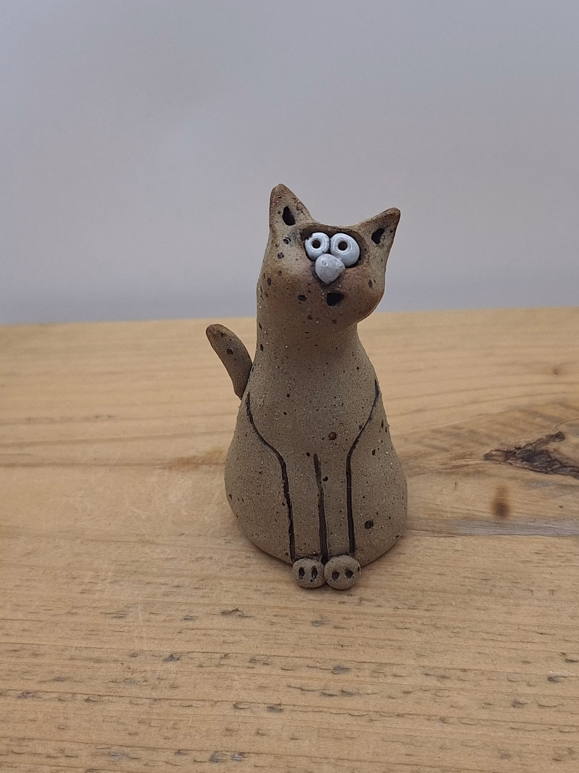 Small ceramic cat figurine with wide painted eyes, sitting upright with paws together, speckled neutral surface and painted whisker details.