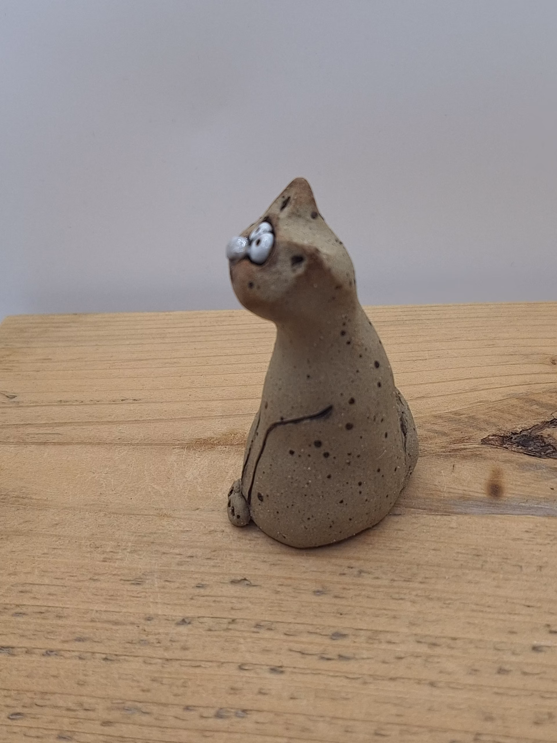 Small ceramic cat figurine with wide painted eyes, sitting upright with paws together, speckled neutral surface and painted whisker details.
