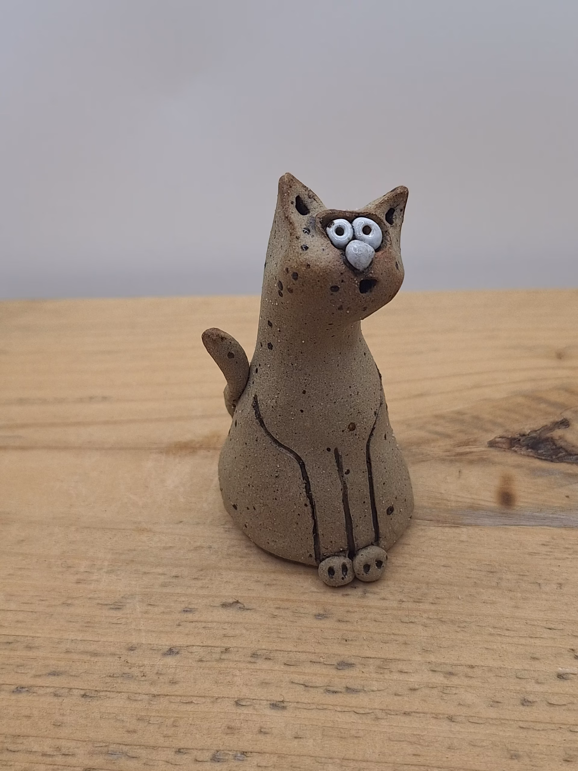 Small ceramic cat figurine with wide painted eyes, sitting upright with paws together, speckled neutral surface and painted whisker details.
