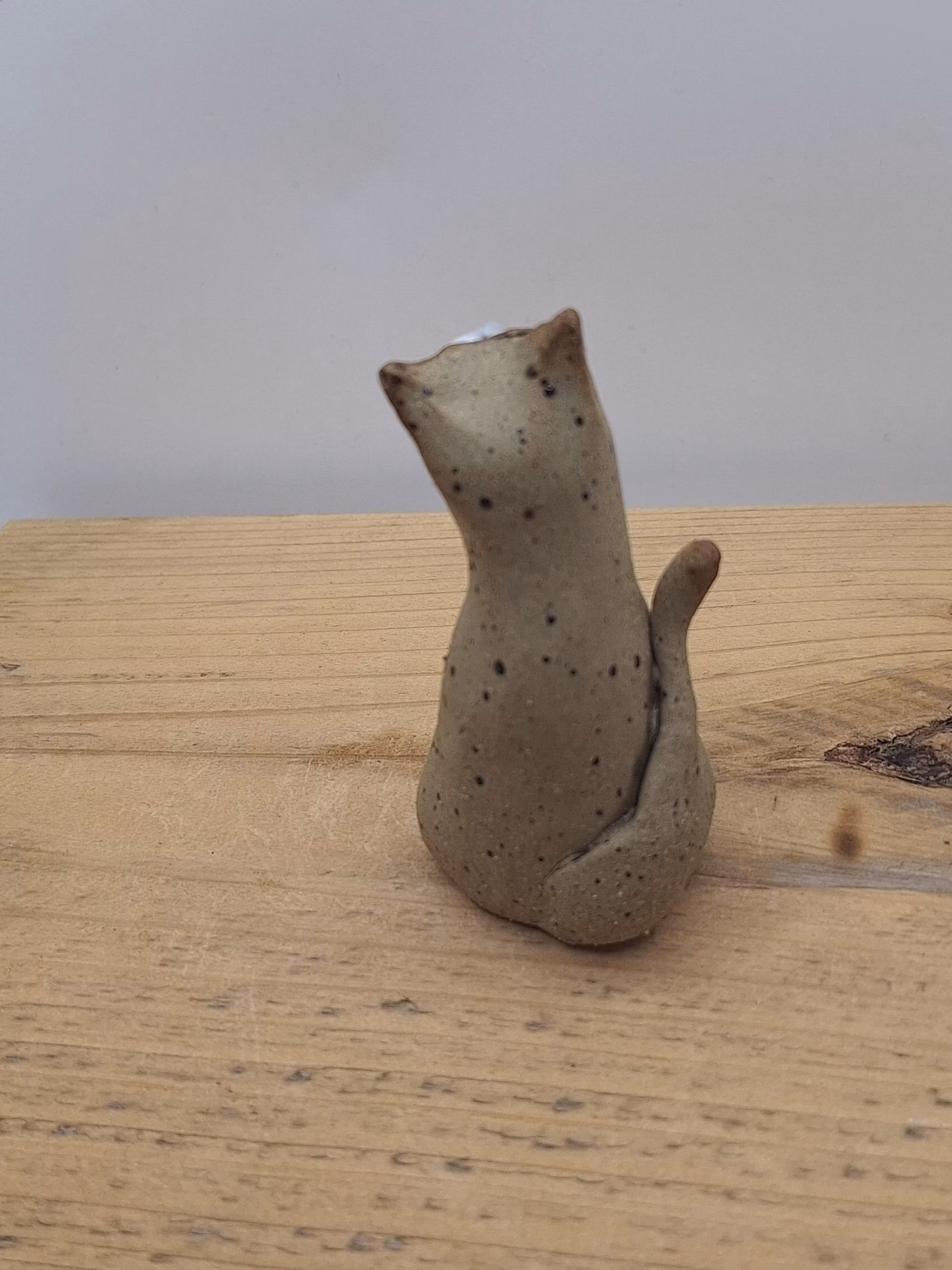 Small ceramic cat figurine with wide painted eyes, sitting upright with paws together, speckled neutral surface and painted whisker details.