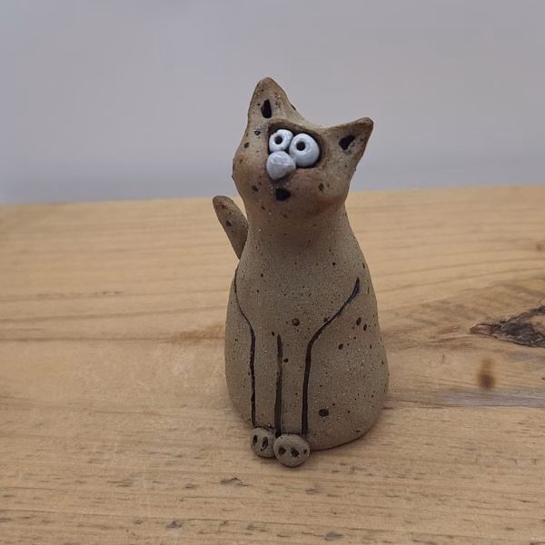 Small ceramic cat figurine with wide painted eyes, sitting upright with paws together, speckled neutral surface and painted whisker details.