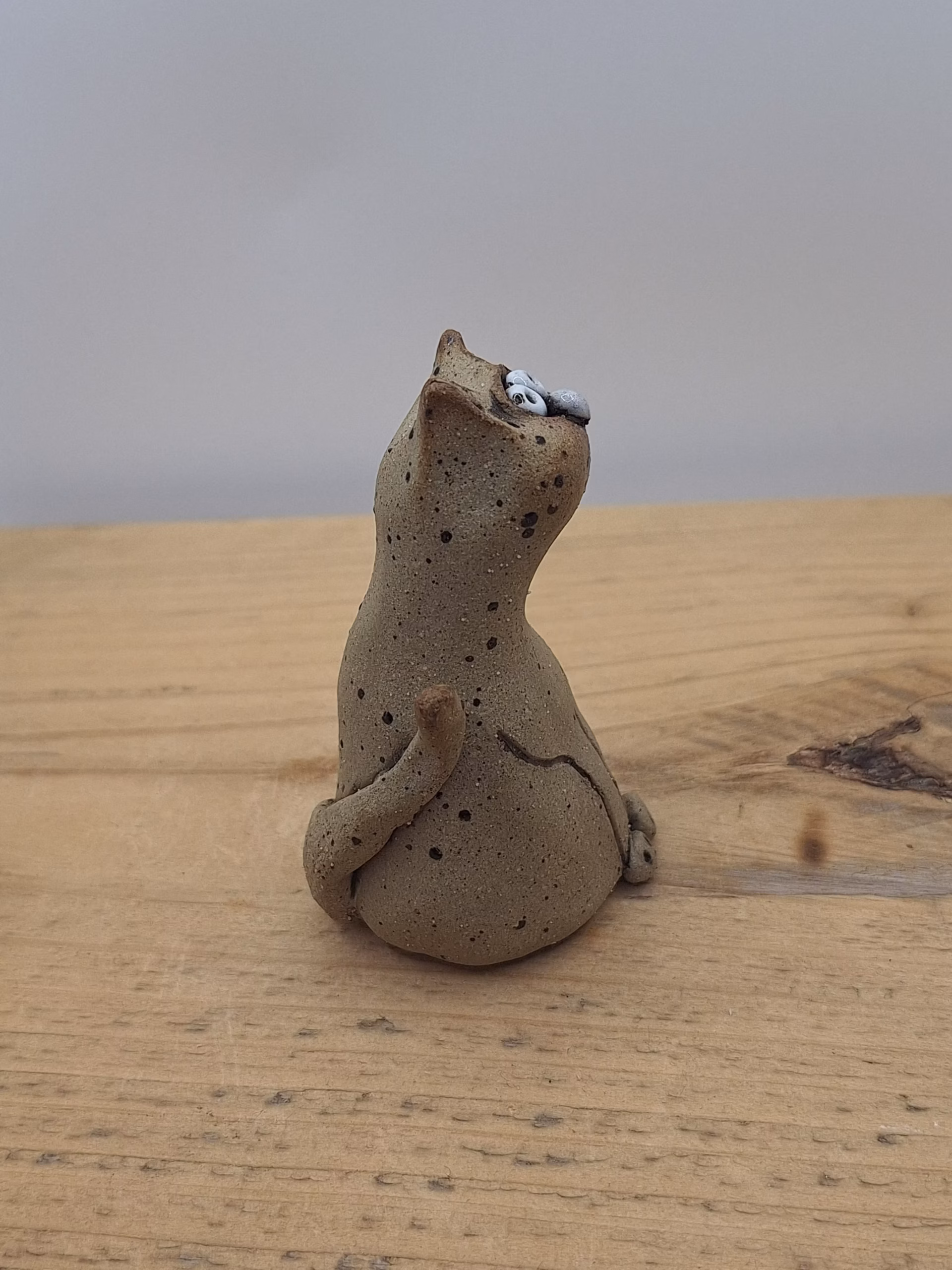 Small ceramic cat figurine with wide painted eyes, sitting upright with paws together, speckled neutral surface and painted whisker details.
