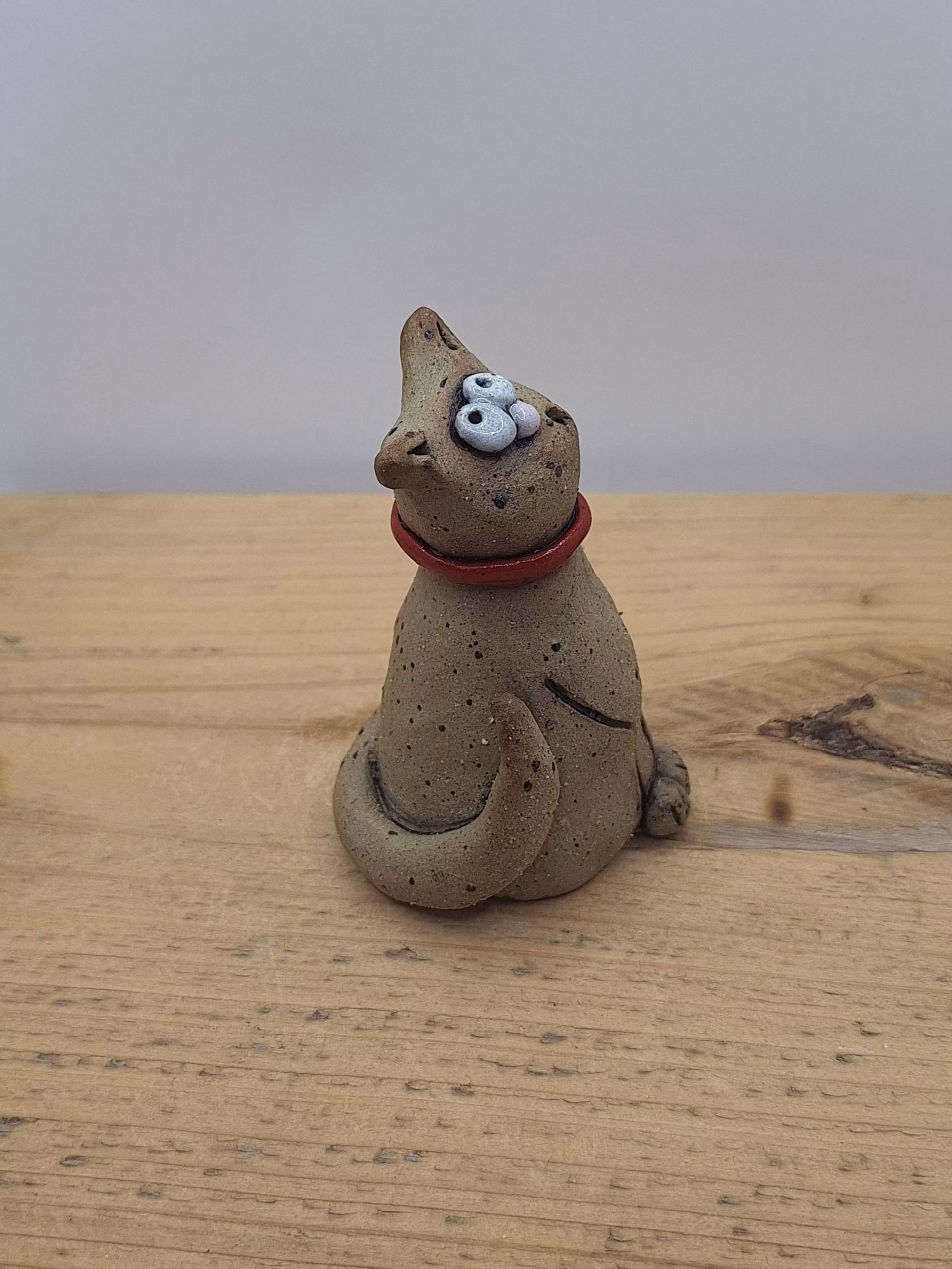 Small ceramic cat with bulging eyes, speckled grey body, red collar with bell, and alert upright ears.