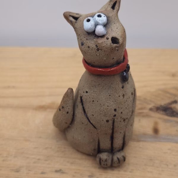 Small ceramic cat with bulging eyes, speckled grey body, red collar with bell, and alert upright ears.