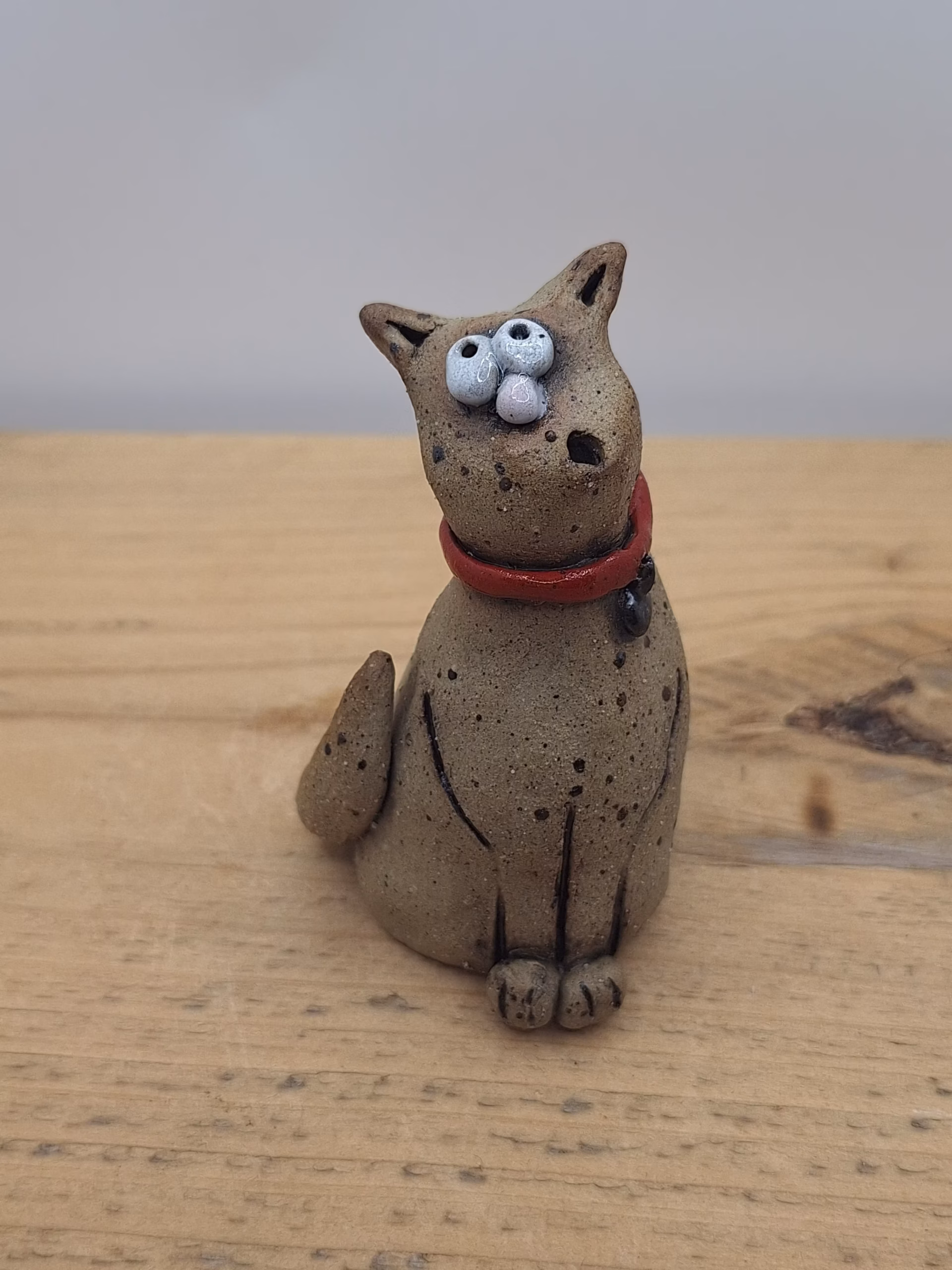Small ceramic cat with bulging eyes, speckled grey body, red collar with bell, and alert upright ears.