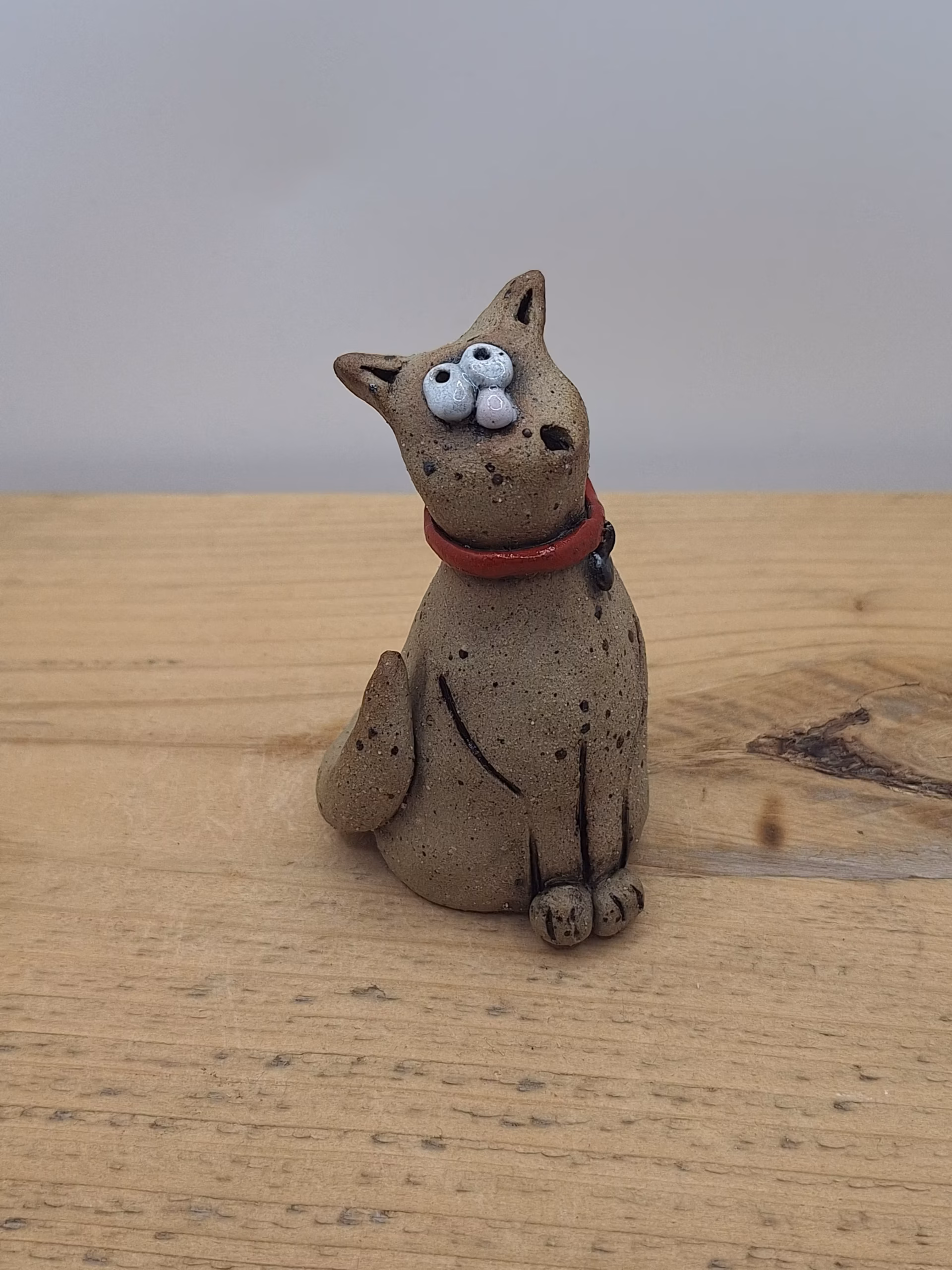 Small ceramic cat with bulging eyes, speckled grey body, red collar with bell, and alert upright ears.