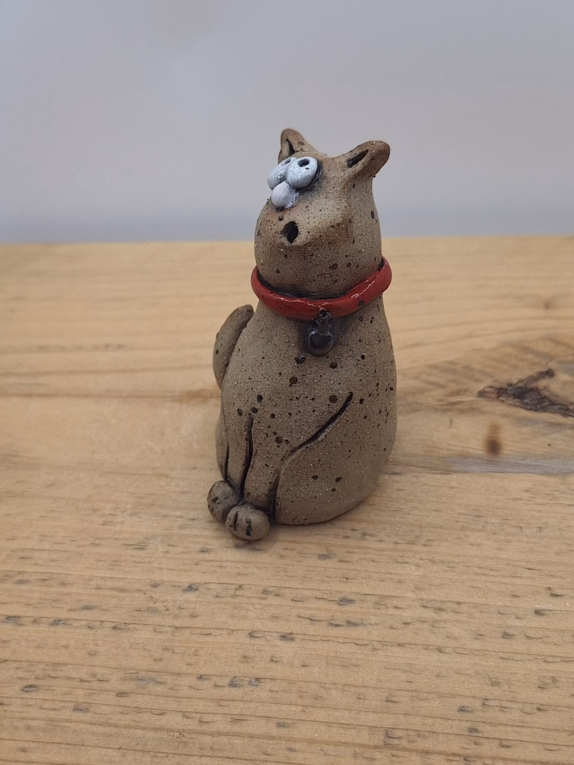 Small ceramic cat with bulging eyes, speckled grey body, red collar with bell, and alert upright ears.