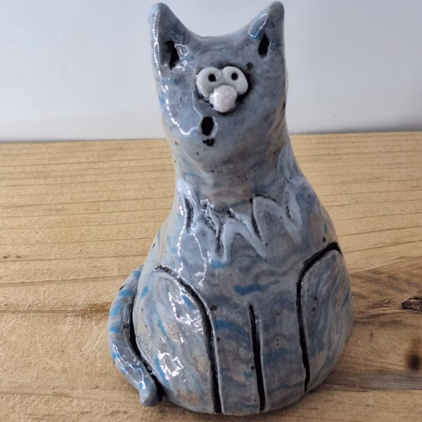Small ceramic cat figurine with grey-blue glaze, white nose and eyes, black pupils and whiskers, seated upright.