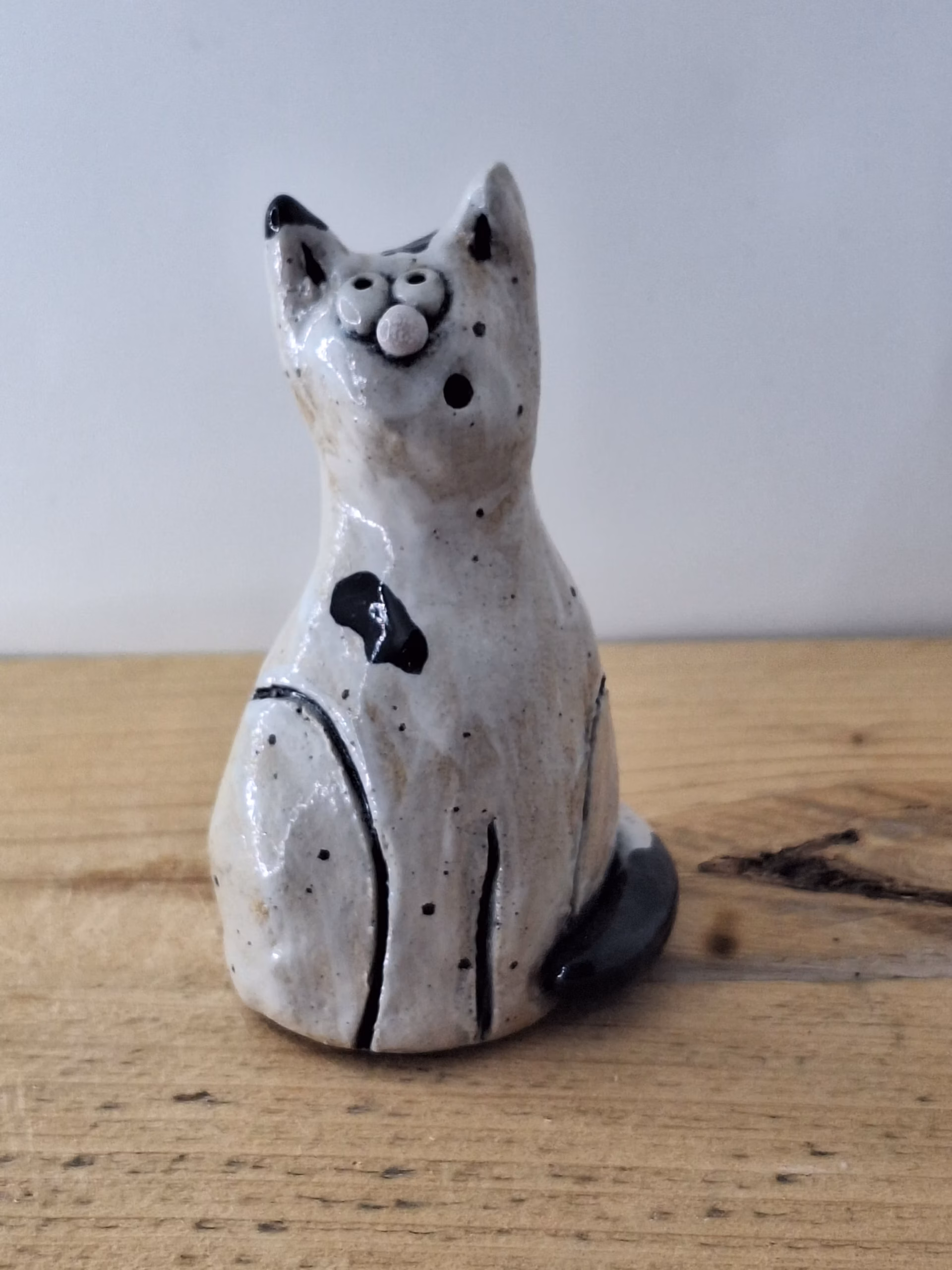 Small ceramic cat figurine with white glaze, black painted details, wide eyes and pink nose, sitting upright.