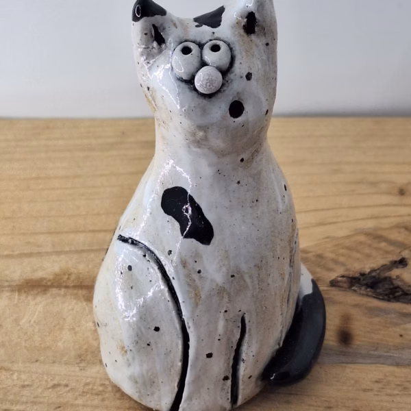 Small ceramic cat figurine with white glaze, black painted details, wide eyes and pink nose, sitting upright.