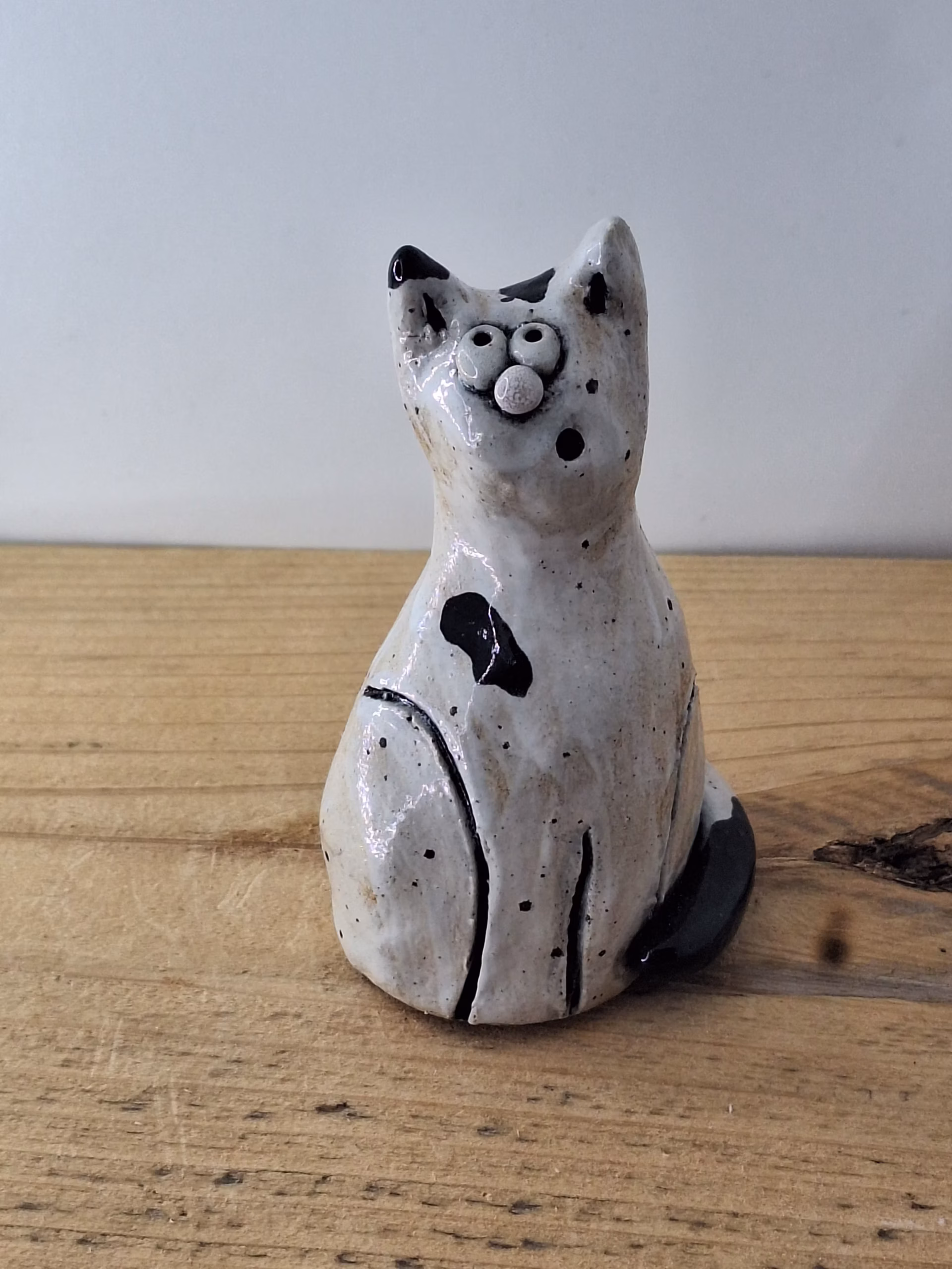 Small ceramic cat figurine with white glaze, black painted details, wide eyes and pink nose, sitting upright.