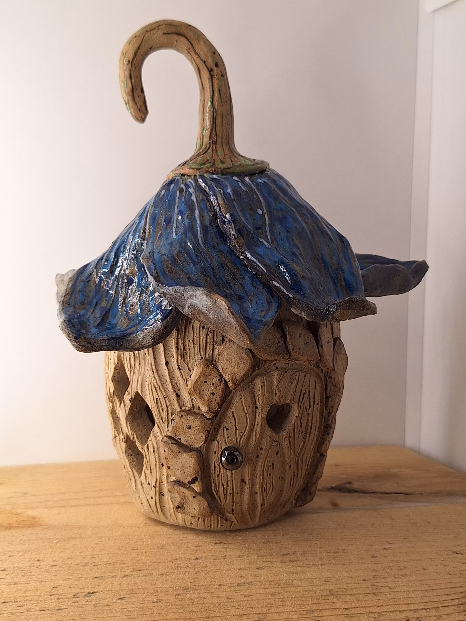 Small ceramic fairy house shaped like a bulbous acorn with deep wood-grain texture, deep blue draped fabric, curved hook stem, and a small heart-shaped door.