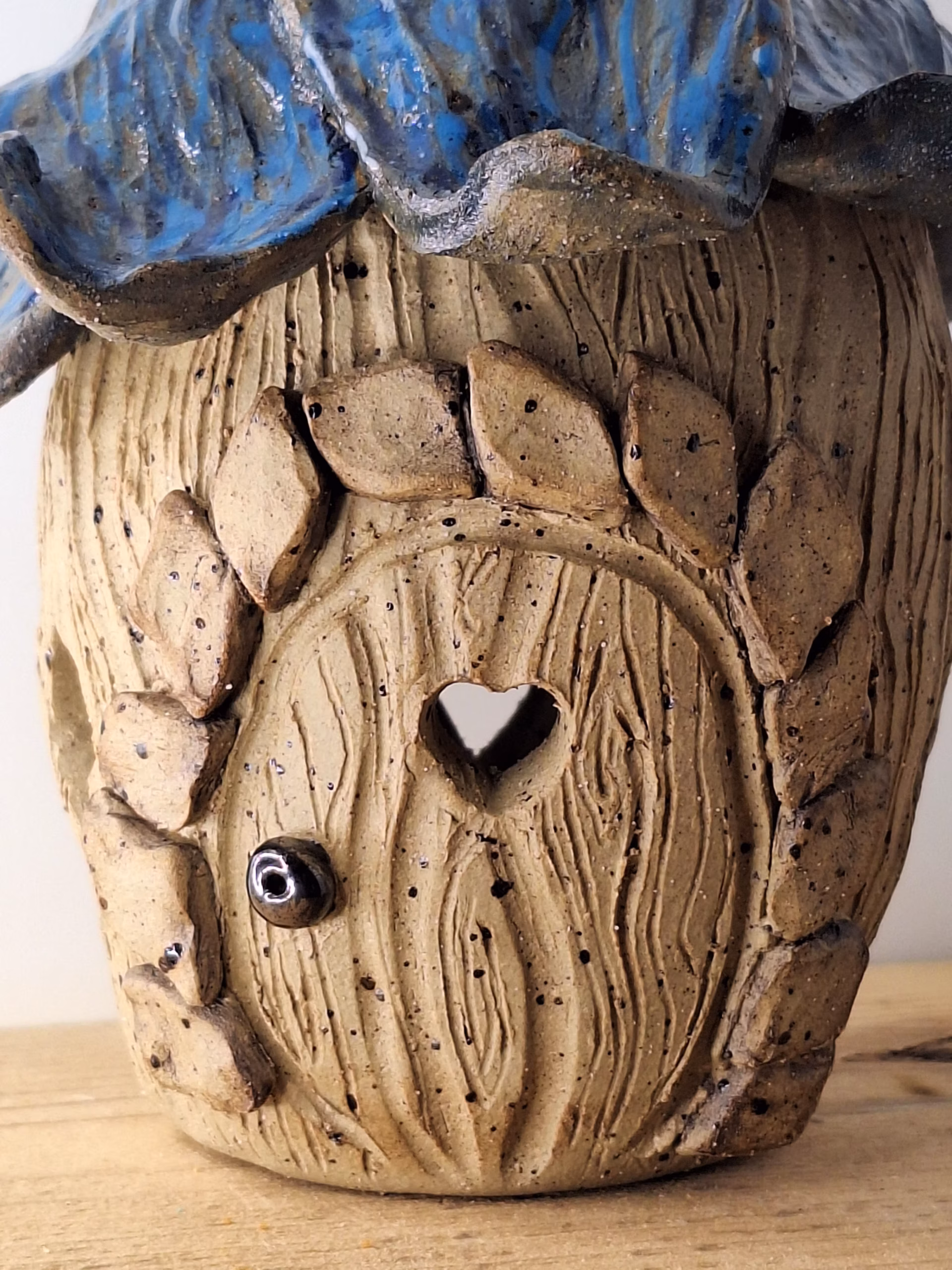 Small ceramic fairy house shaped like a bulbous acorn with deep wood-grain texture, deep blue draped fabric, curved hook stem, and a small heart-shaped door.
