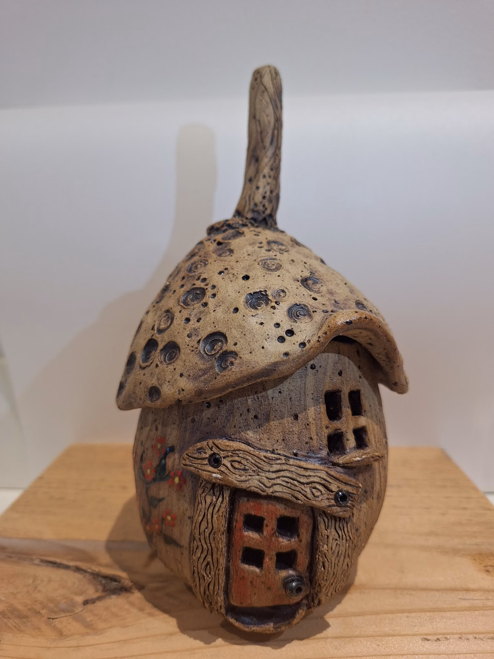 Small ceramic acorn house with a textured roof, wooden branch door frame, and red-painted lower storey with four square windows.