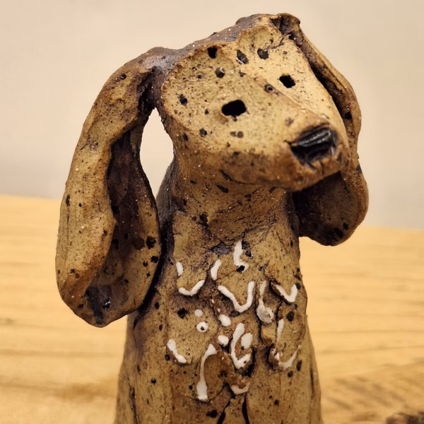 Small ceramic dog figurine with long ears, speckled brown glaze, and white painted markings on chest.