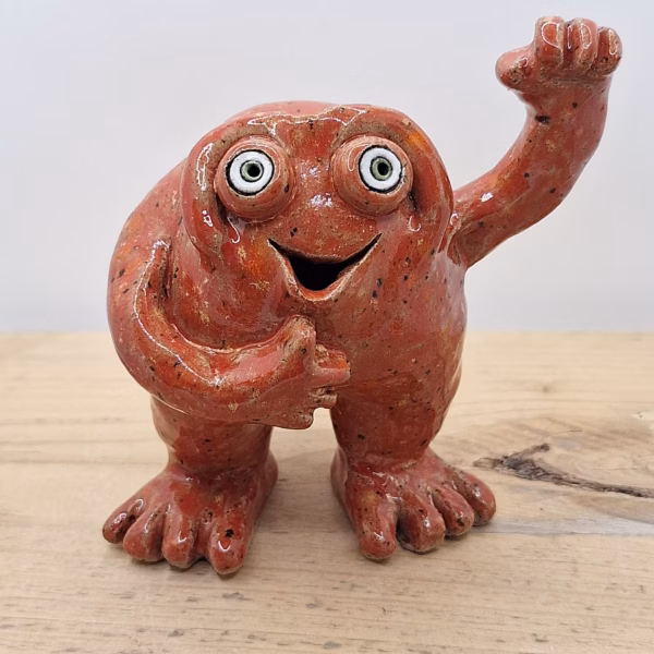 Lennard, a cheerful red ceramic monster with wide eyes, raised fist and open-mouthed grin, standing on splayed feet.