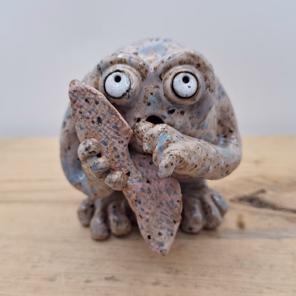 Small ceramic monster with wide round eyes, textured speckled surface, and a leaf-like wing held to its mouth in a surprised expression.