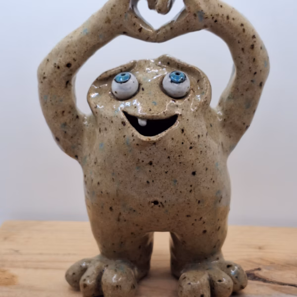 Small ceramic monster with blue eyes, wide grin and chunky feet, arms raised in a heart shape above its head.