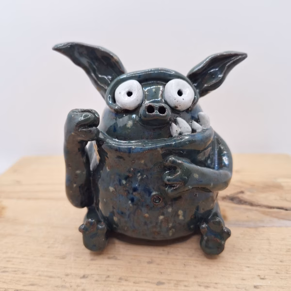 Small dark ceramic monster with large round white eyes, pointed ears, stubby limbs and a wide grin.