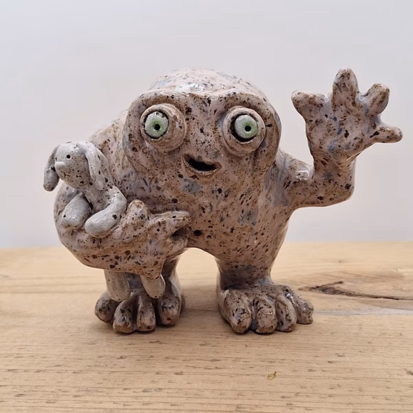 Small ceramic monster with wide eyes, open mouth smile, textured speckled surface, holding a tiny rabbit, one hand raised in greeting.